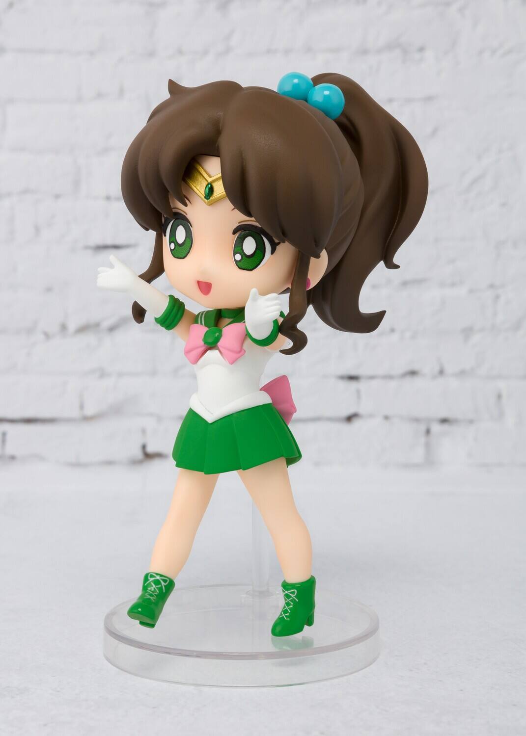 The image shows a figurine of a character with long brown hair tied with blue hair accessories. She is wearing a white top with a green collar and a pink bow, along with a green skirt. The character is also wearing green boots and is posed with her hands raised.