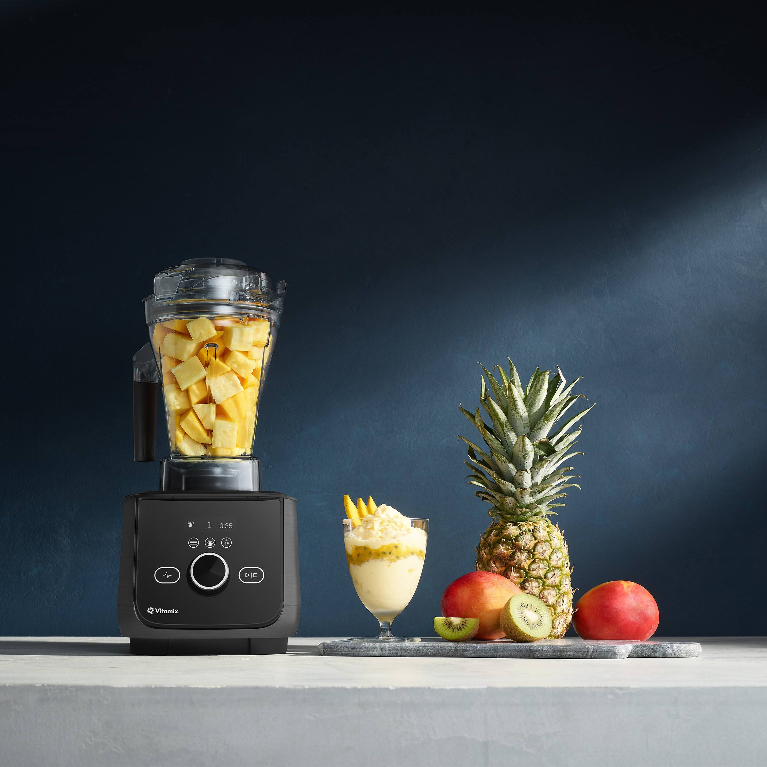 The image features a blender filled with fruit, including apples and oranges, along with a pineapple. The blender is placed on a table, and there is a glass of the blended fruit nearby. The scene is set against a dark blue background, which adds a dramatic touch to the image. The blender is a prominent feature in the scene, and the assortment of fruits creates a colorful and appetizing display.