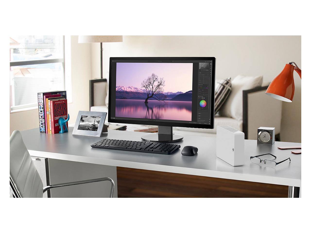The photo shows a modern workspace with a computer monitor displaying a serene landscape. On the desk, there are several books, including "The Photographer's Eye" and "David Ogilvy on Advertising." There's also a framed photograph, a small blue figurine, a red desk lamp, a clock, a pair of glasses, and a white computer tower.