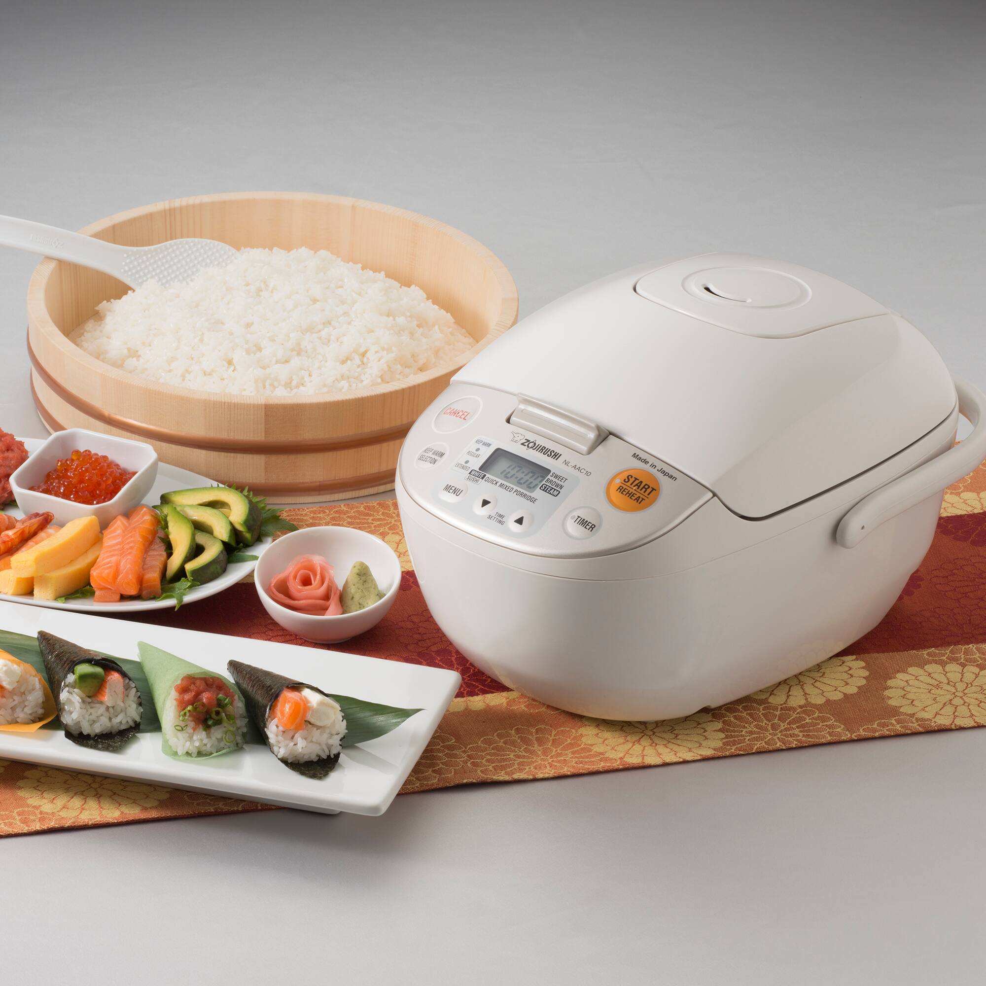 The image features a variety of food items, including a bowl of rice, a bowl of sushi, and a bowl of vegetables. There is also a white rice cooker on the table, which is likely used for preparing the rice. The food items are arranged on a dining table, creating an appetizing display.