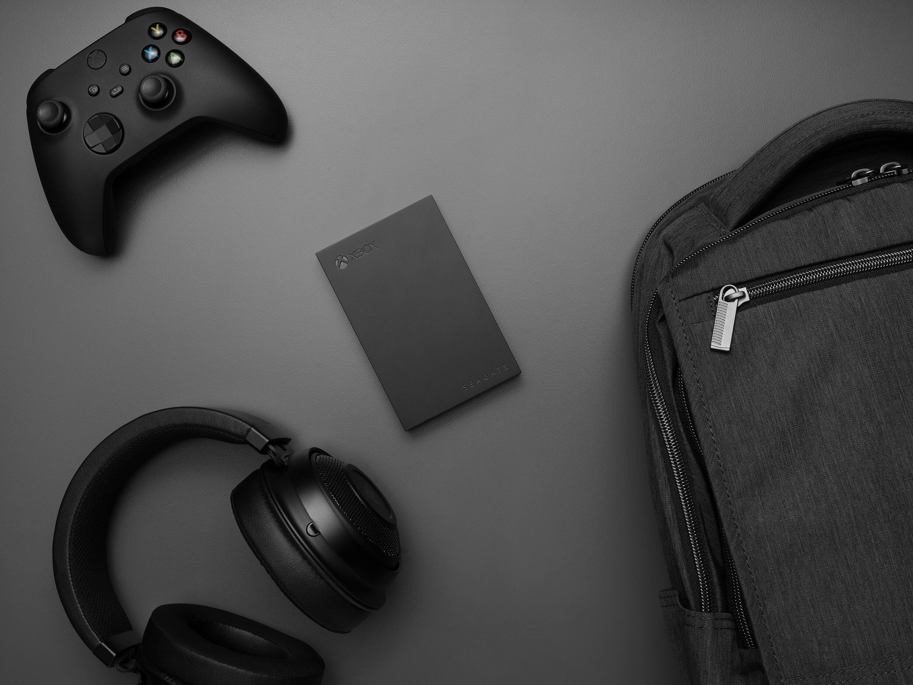 The image features a black Xbox controller, a cell phone, and a backpack. The Xbox controller is placed next to the cell phone, which is positioned near the backpack. The backpack is located on the right side of the image, and the cell phone is situated in the middle.