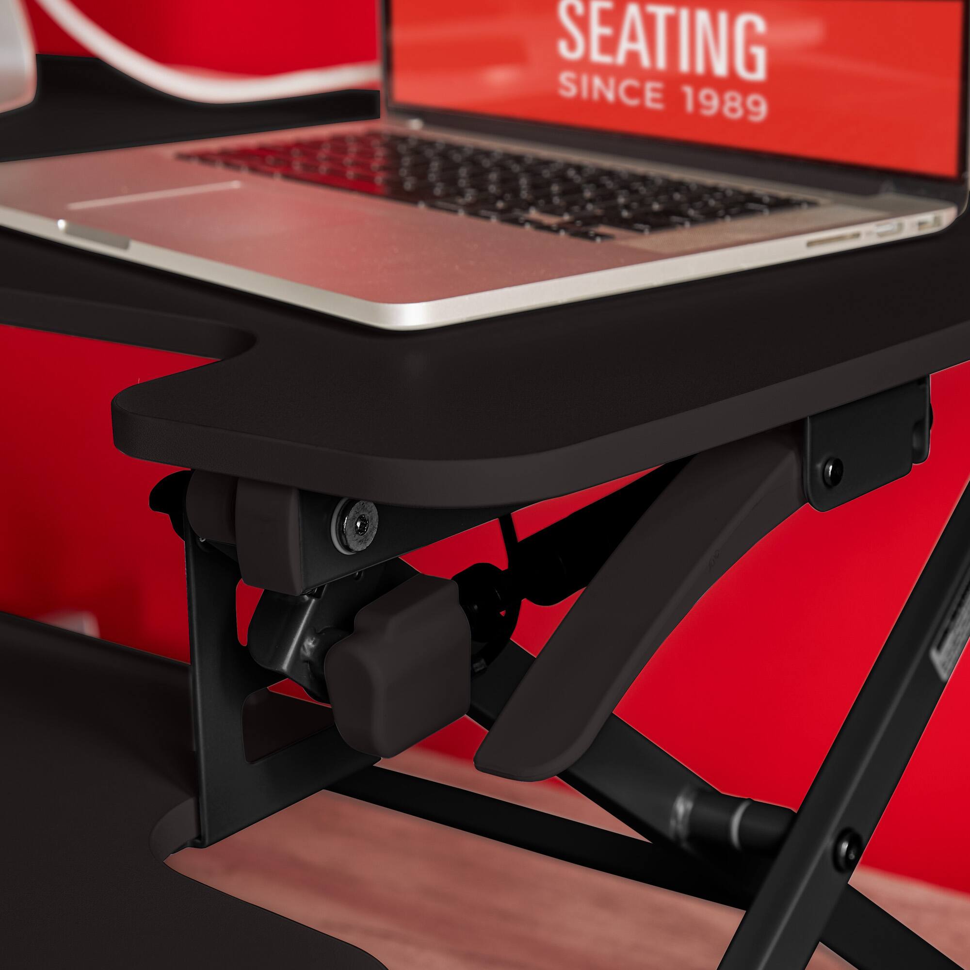 The image features a laptop computer sitting on a black stand or a laptop stand, which is placed on a desk. The laptop stand is designed to elevate the laptop to a more comfortable viewing angle, providing better ergonomics for the user. The laptop stand is positioned in front of a red wall, which adds a pop of color to the scene. The image does not provide any information about the brand or model of the laptop.