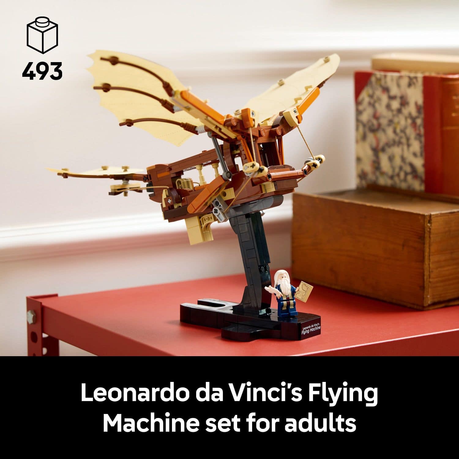 The image features a red table with a model of Leonardo da Vinci's Flying Machine set for adults. The model is made of Lego bricks and is displayed on the table. The table also has a book on it, adding to the overall scene.