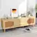 The image features a wooden dresser or desk with a picture on the wall above it. The dresser has a drawer and a shelf, and it is placed on a carpet. On the dresser, there are several books, a vase, and a clock. Additionally, there is a potted plant nearby, and a robotic vacuum cleaner is present on the floor. The scene appears to be a well-organized and functional living space.