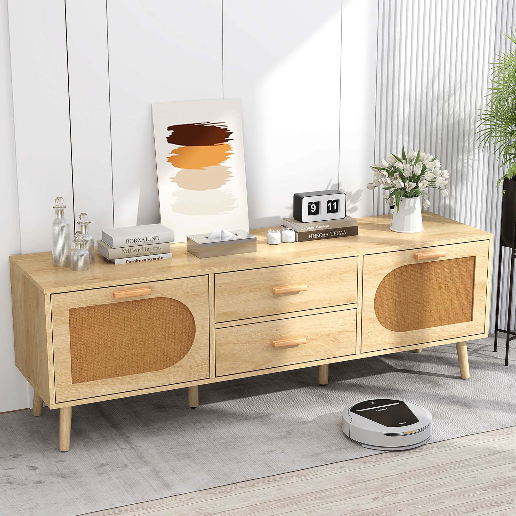 The image features a wooden dresser or desk with a picture on the wall above it. The dresser has a drawer and a shelf, and it is placed on a carpet. On the dresser, there are several books, a vase, and a clock. Additionally, there is a potted plant nearby, and a robotic vacuum cleaner is present on the floor. The scene appears to be a well-organized and functional living space.