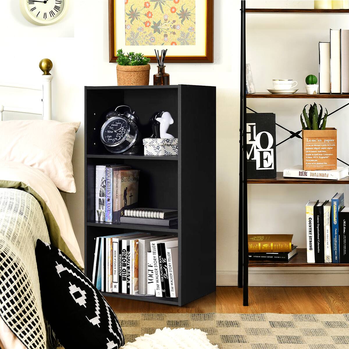 The image shows a bedroom with a black bookshelf filled with books. The shelf is placed next to a bed, and there is a clock on the shelf as well. The room also features a potted plant and a vase, adding to the cozy atmosphere. The books on the shelf are arranged in a way that makes them easily accessible, and the variety of books suggests that the person living in this space enjoys reading and has diverse interests.