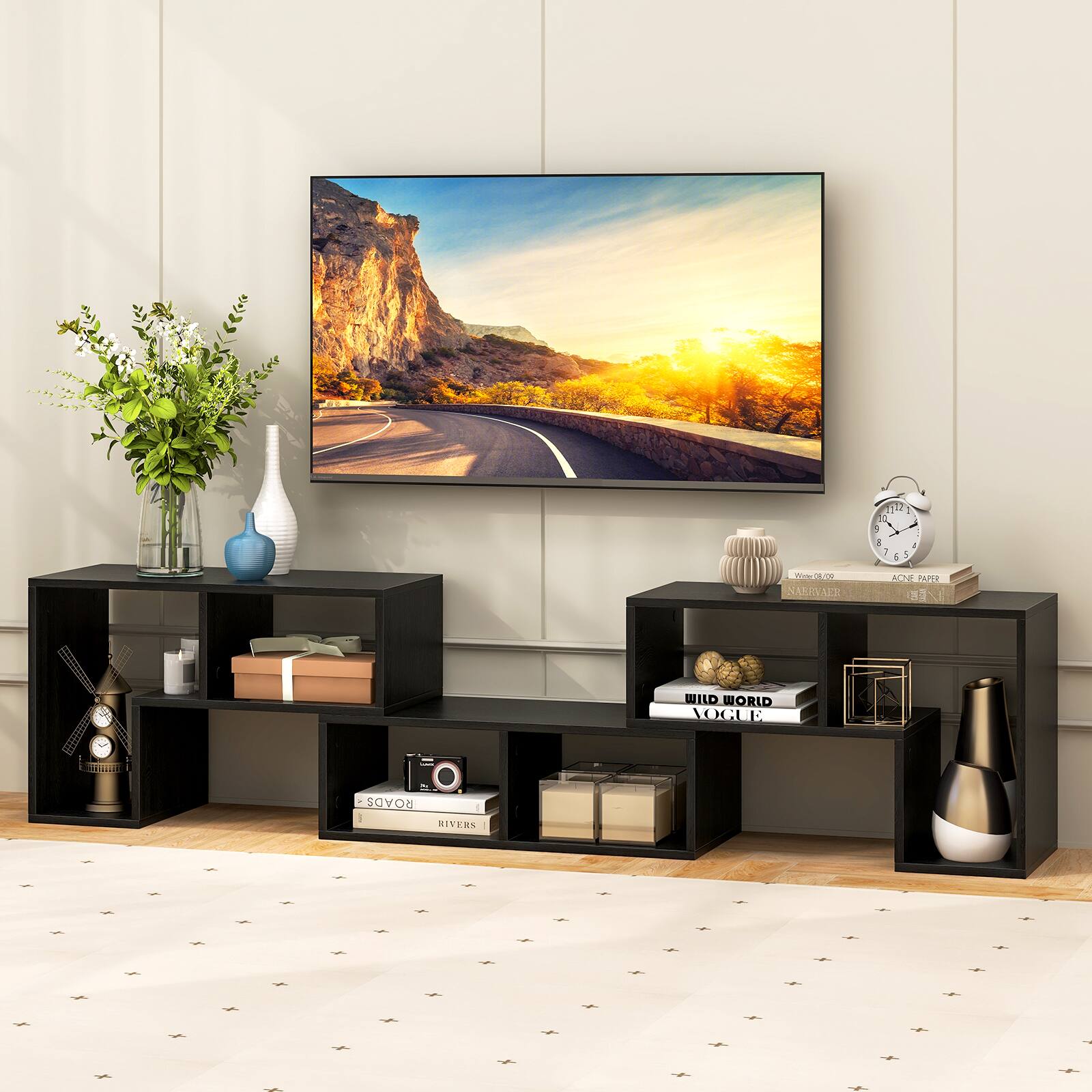 A large flat screen TV is mounted on the wall. The TV is displaying a picture of a road surrounded by mountains. The television is placed above a black table, which has a vase, a clock, and a book on it. The table is situated in front of a window, and there is a potted plant nearby.