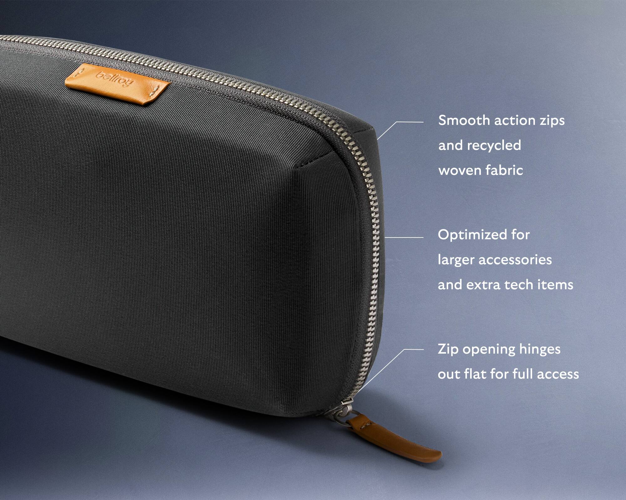 The image features a black bag with a zipper and a tag that says "Smooth action zips and recycled woven fabric." The tag also provides information about the bag's features, such as the optimized design for larger accessories and extra tech items, the zip opening hinges that allow the bag to open flat for full access, and the zipper's smooth action. The bag is displayed on a blue background, and the zipper is open, revealing the interior of the bag.