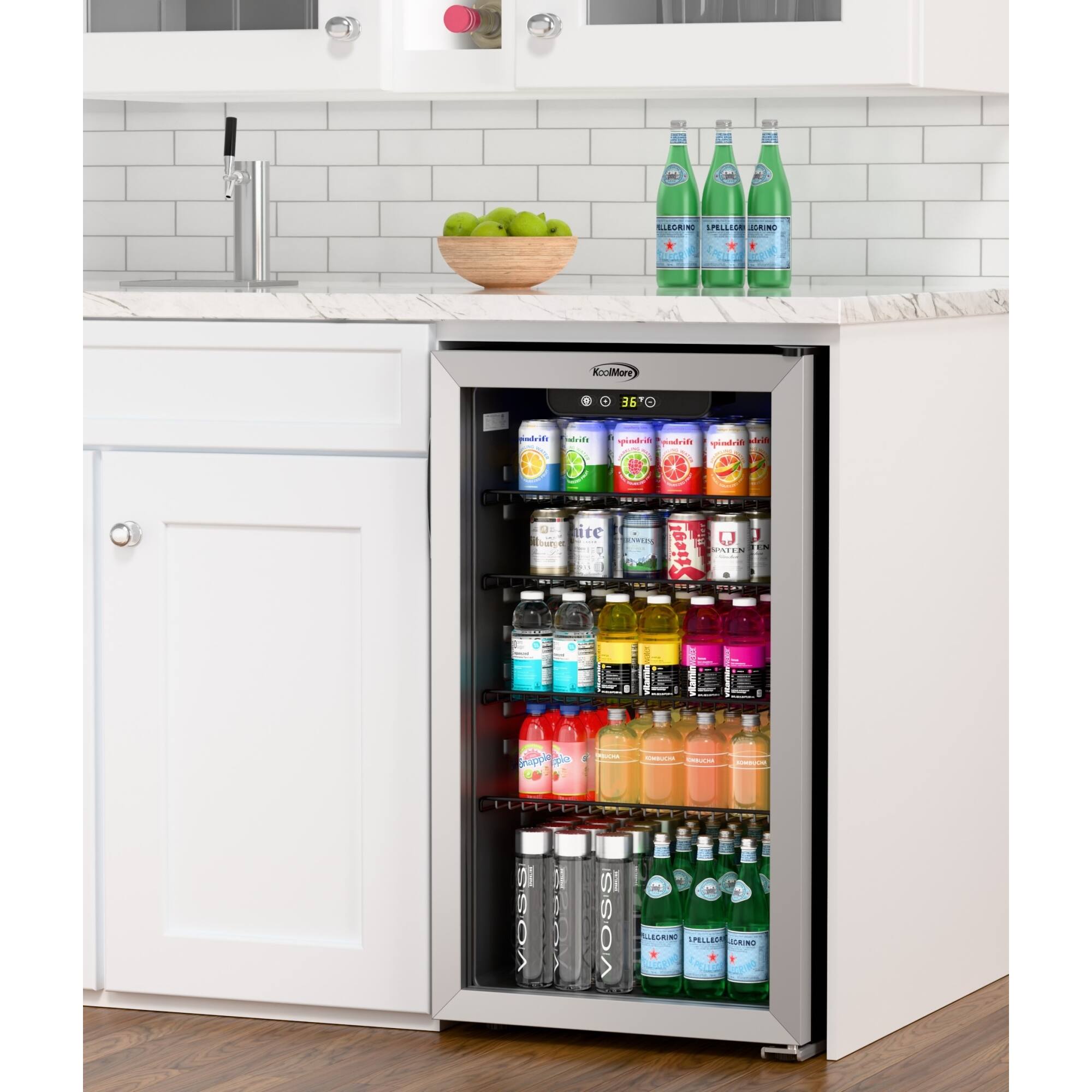 The image shows a kitchen with a refrigerator that has a drink dispenser on the door. The refrigerator is stocked with a variety of drinks, including bottles of water, soda, and juice. There are also some apples placed on the countertop. The kitchen appears to be well-organized and ready for use.