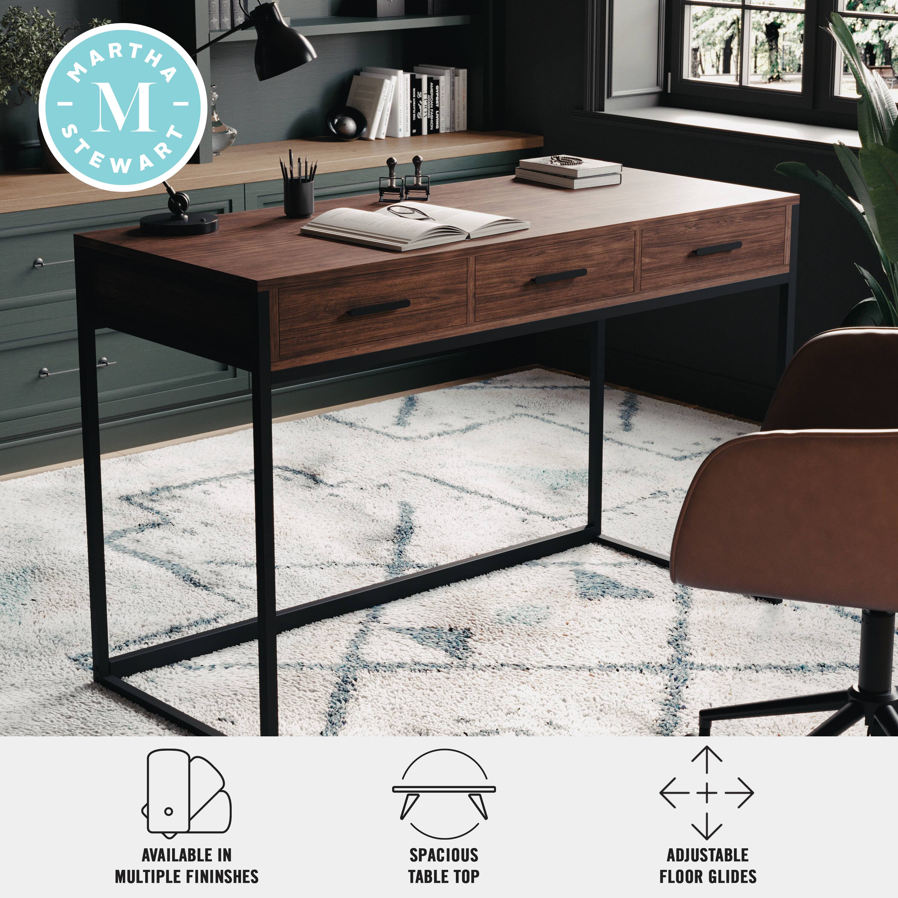 The image features a Martha Stewart desk with a wooden top and a black metal base. The desk is placed on a rug and has a chair positioned in front of it. The desk is equipped with a lamp and a book, and there are several potted plants nearby. The desk is available in multiple finishes and comes with adjustable floor glides for added stability and comfort.