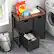 The image features a laundry room with a washing machine and a dryer. There is a small table with a basket and a few items on it, including a bottle of detergent. The table is located near a window, and there is a trash can nearby. The laundry room also has a shelf with a few items on it, such as a towel and a book.