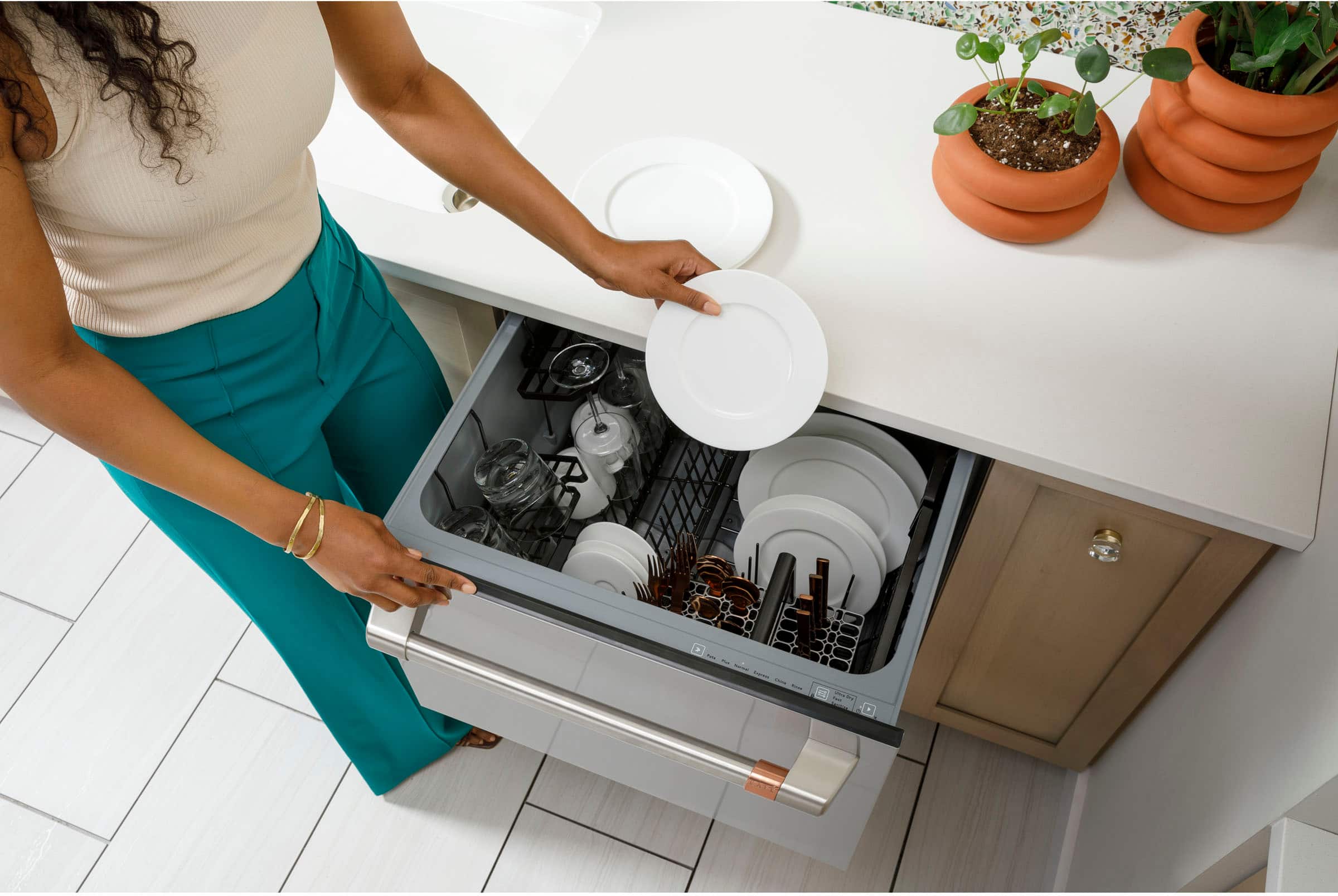 The image shows a woman standing in a kitchen, holding a plate over a dishwasher. She is placing the plate into the dishwasher, which is located under the kitchen counter. The kitchen counter has several potted plants on it, adding a touch of greenery to the space. The woman is wearing a white shirt and green pants, and she is focused on her task of loading the dishwasher.