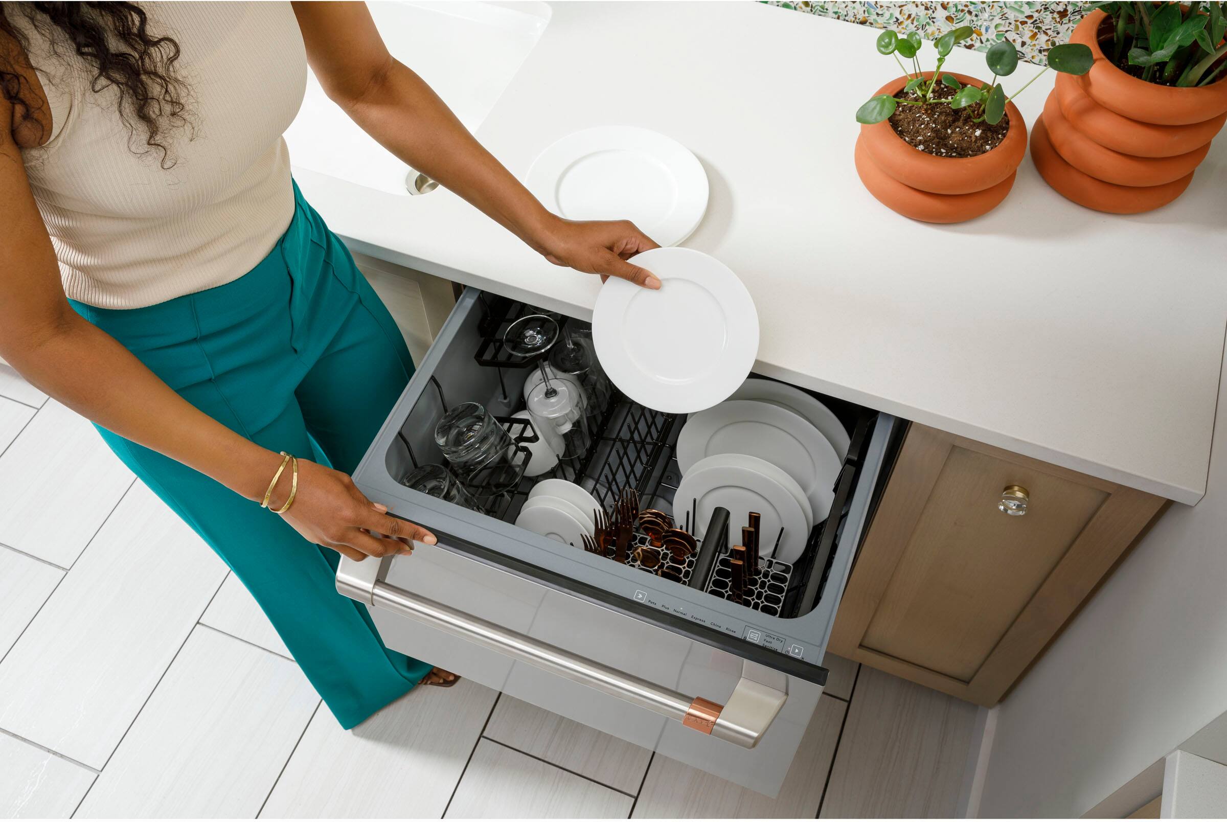 The image shows a woman standing in a kitchen, holding a plate over a dishwasher. She is placing the plate into the dishwasher, which is located under the kitchen counter. The kitchen counter has several potted plants on it, adding a touch of greenery to the space. The woman is wearing a white shirt and green pants, and she is focused on her task of loading the dishwasher.