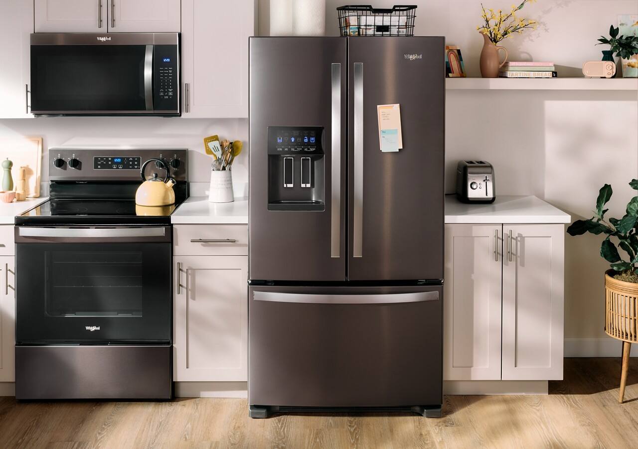 The image shows a kitchen with a refrigerator, microwave, oven, and stove. The refrigerator is a large black and silver model, and it is placed next to a microwave oven. The oven is located below the microwave, and the stove is situated to the left of the refrigerator. The kitchen also features a sink and a potted plant, adding a touch of greenery to the space.