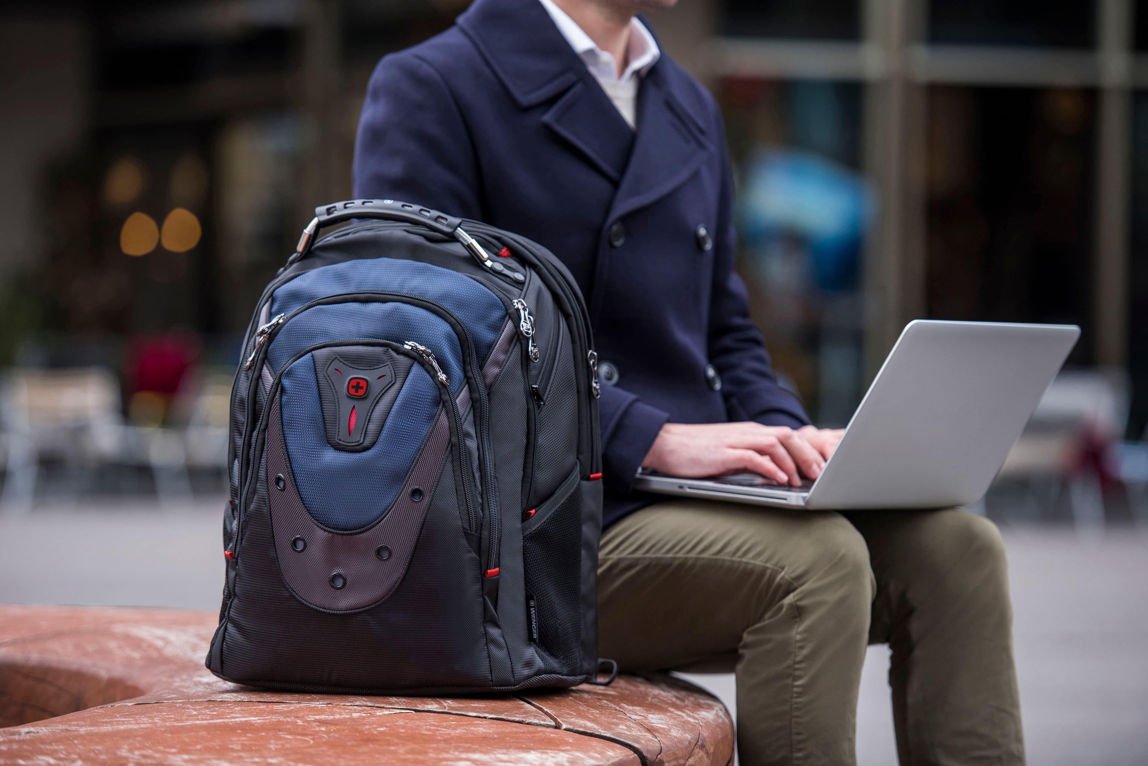 The man is sitting on a bench with a laptop and a backpack.