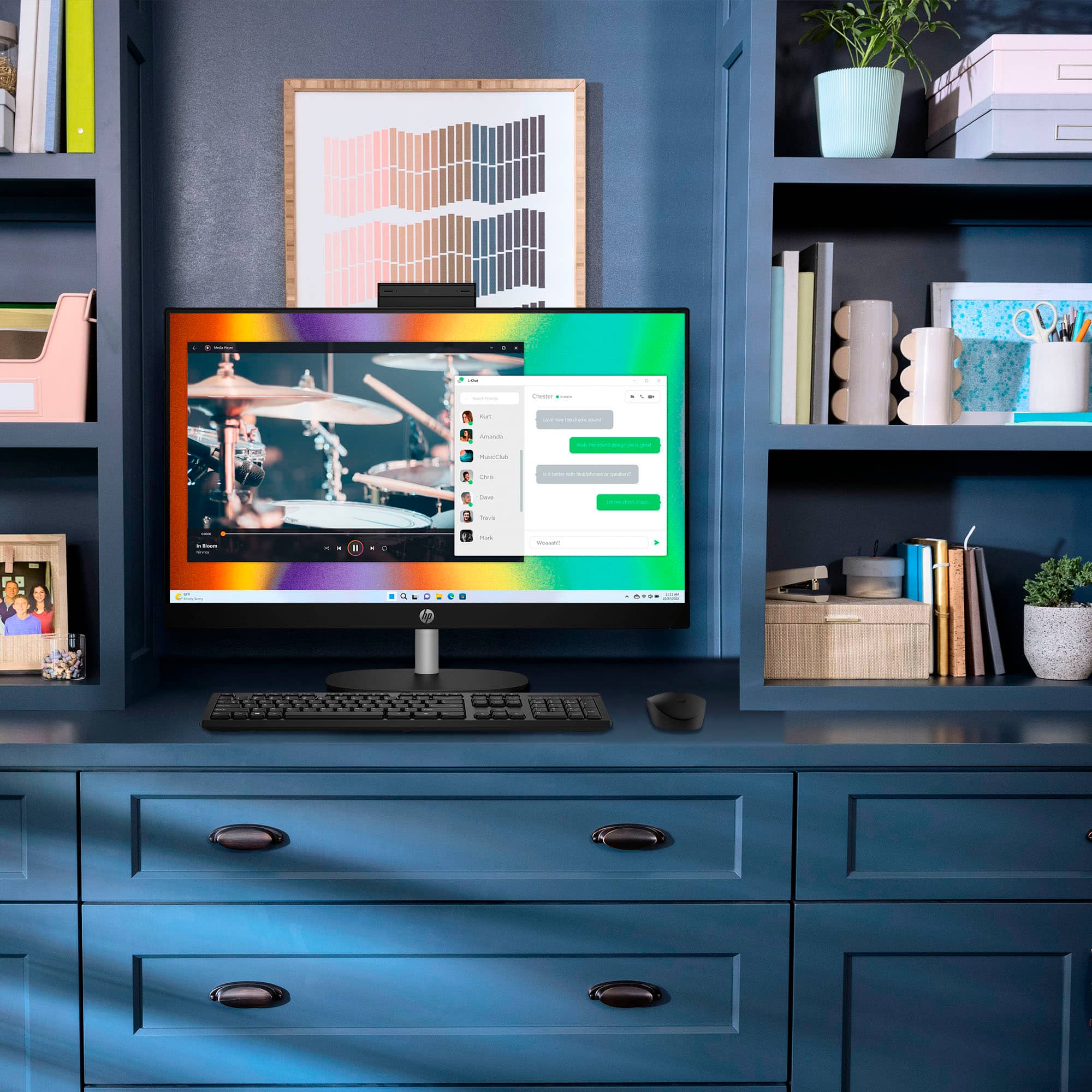 The image features a computer desk with a desktop computer, keyboard, and mouse. The computer screen displays a chat window, and there are several books nearby. The desk is situated in front of a blue wall, and there is a potted plant nearby. Additionally, there is a vase on the desk, and a cup can be seen in the background. The scene is likely a home office or workspace.
