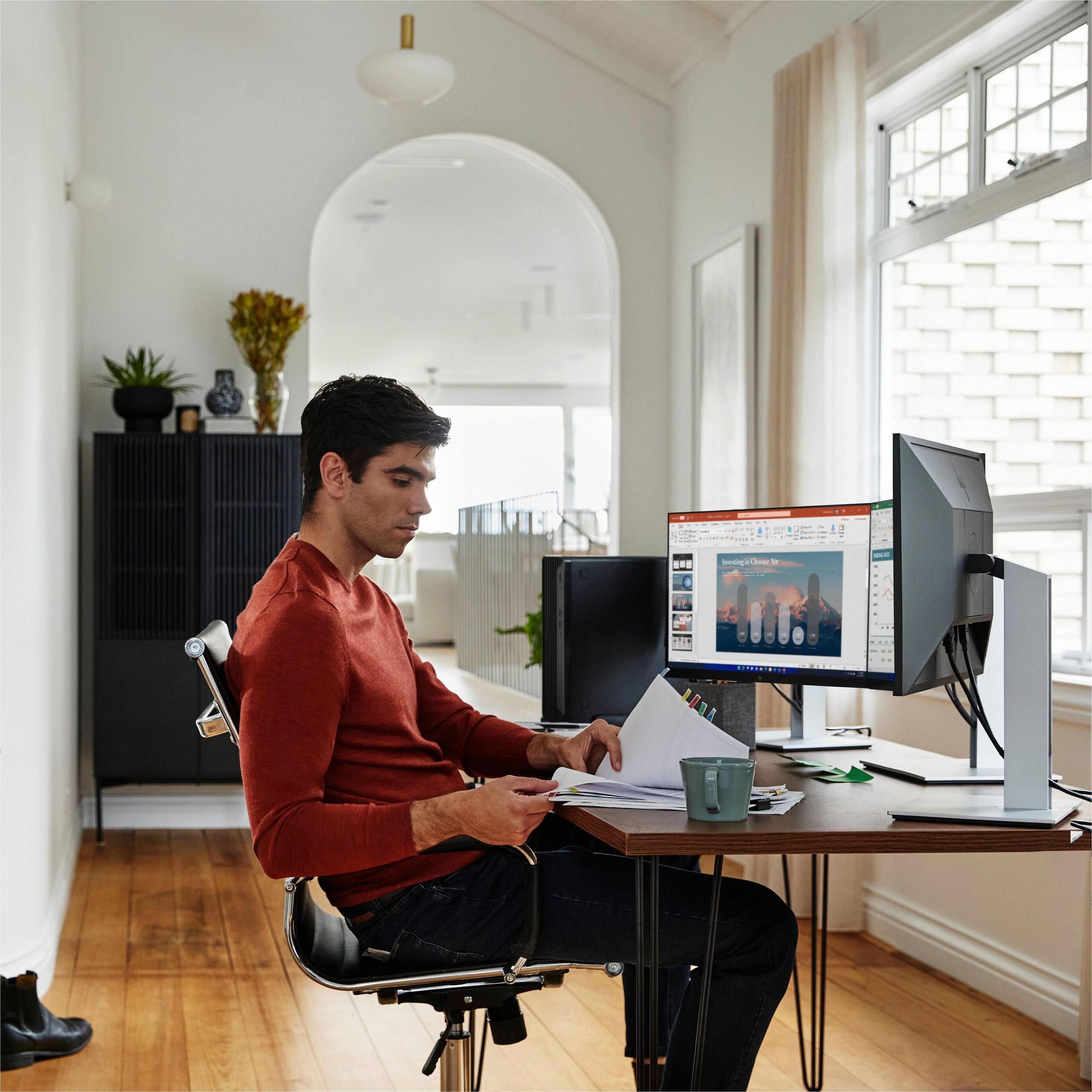 The man is sitting at a desk with a computer monitor, a keyboard, and a mouse. He is wearing a red shirt and is looking at the screen. There is a cup on the desk, and a potted plant is nearby. The man is also holding a book, possibly reading or studying.