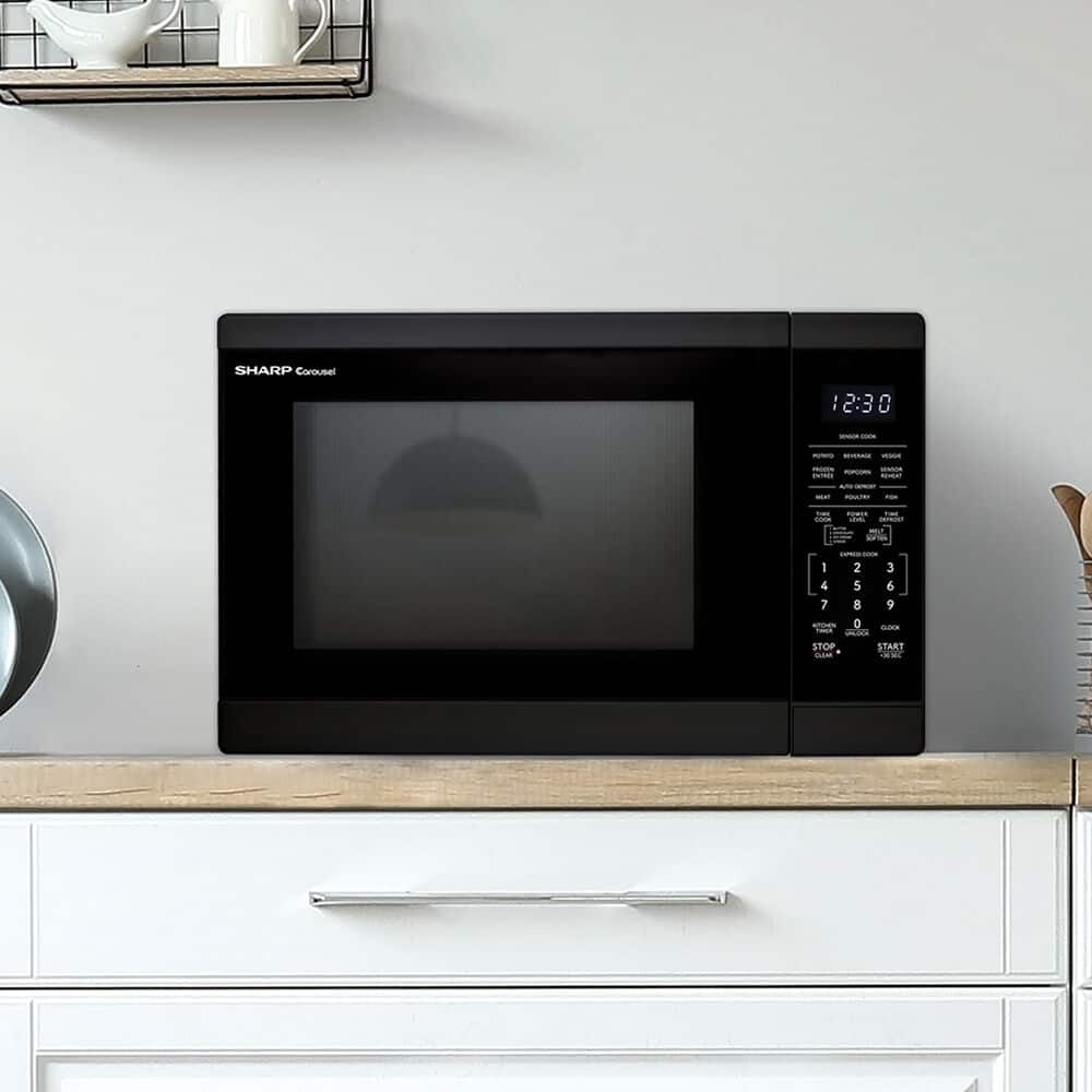 The image shows a black Sharp microwave sitting on top of a white counter. The microwave is currently displaying the time of 12:30.