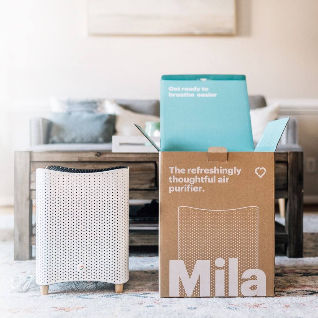 The image features a box with the word "Mila" on it, placed on a carpeted floor. Next to the box, there is a small white air purifier. The box is open, revealing the air purifier inside. The scene suggests that the air purifier is a new addition to the living space, and it is ready to be set up and used.