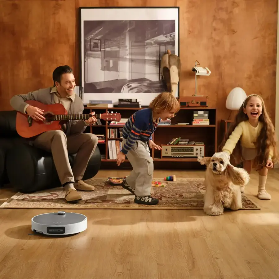 The image shows a man playing a guitar while sitting on a couch, with a young boy and a girl standing in front of him. They are all smiling and enjoying their time together. There is also a dog in the scene, adding to the warm and happy atmosphere. The room features a bookshelf with several books, and a teddy bear is placed nearby. The man is wearing a tie, which adds a touch of formality to the casual setting.