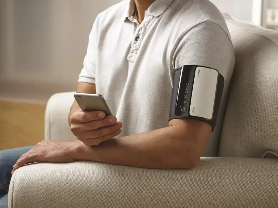 The man is sitting on a couch and holding a cell phone. He is also wearing a device on his arm, possibly a blood pressure monitor.