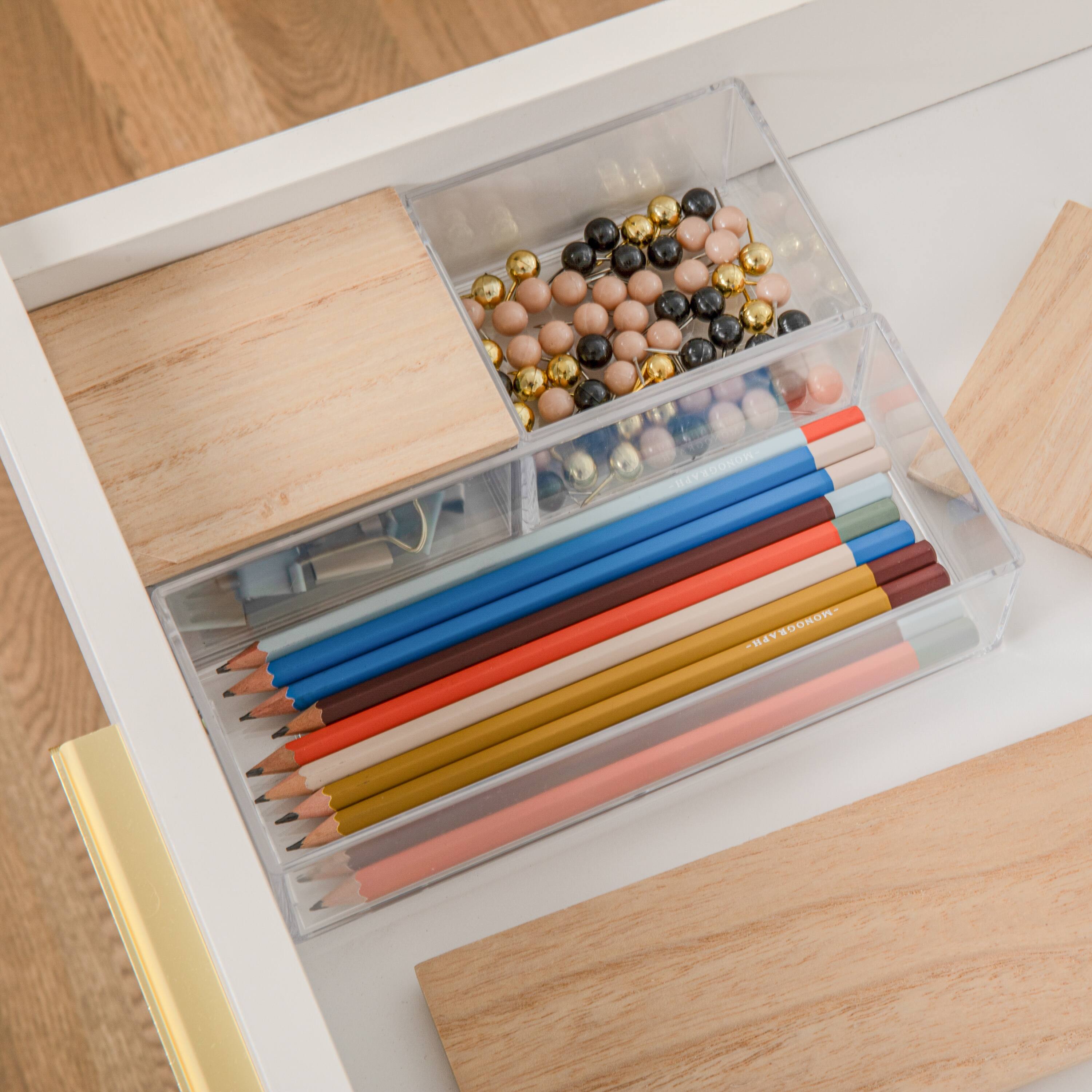 The image shows a wooden desk with a drawer open, revealing a variety of items inside. There are several pencils of different colors, including blue, orange, and red, arranged neatly in the drawer. Additionally, there are two pairs of scissors placed in the drawer, one towards the left side and the other towards the right side. The drawer also contains a small container with beads, adding a touch of personalization to the workspace. The overall scene conveys a well-organized and functional workspace.