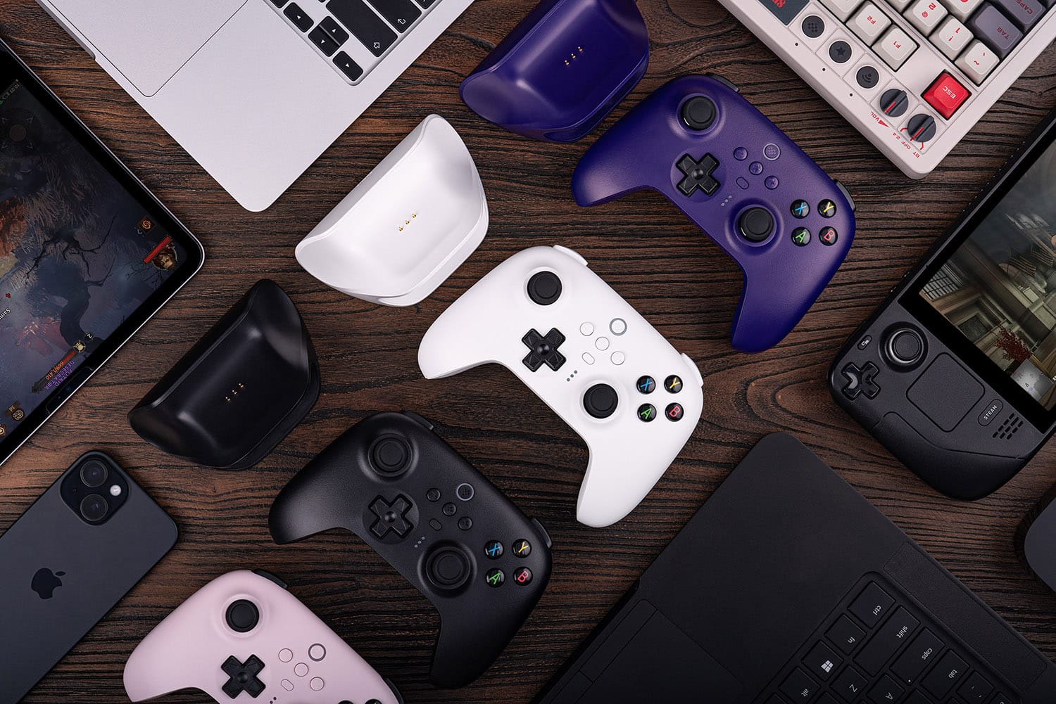 The image shows a table with various electronic devices, including a laptop, a cell phone, a keyboard, and several game controllers. The game controllers are of different colors, including black, white, and purple. The table is also surrounded by other items such as a mouse and a remote control. The arrangement of the devices and items suggests that this is a workspace or a gaming area.