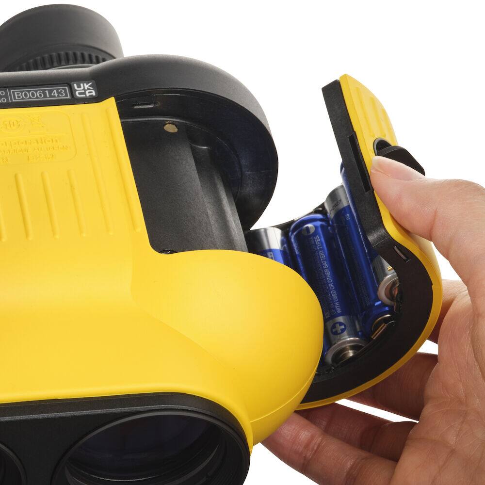 The image shows a person holding a yellow and black device, which appears to be a camera. The person is holding the device in their hand and is in the process of putting batteries into it. The batteries are placed in a compartment on the device, and the person is using their fingers to insert them.