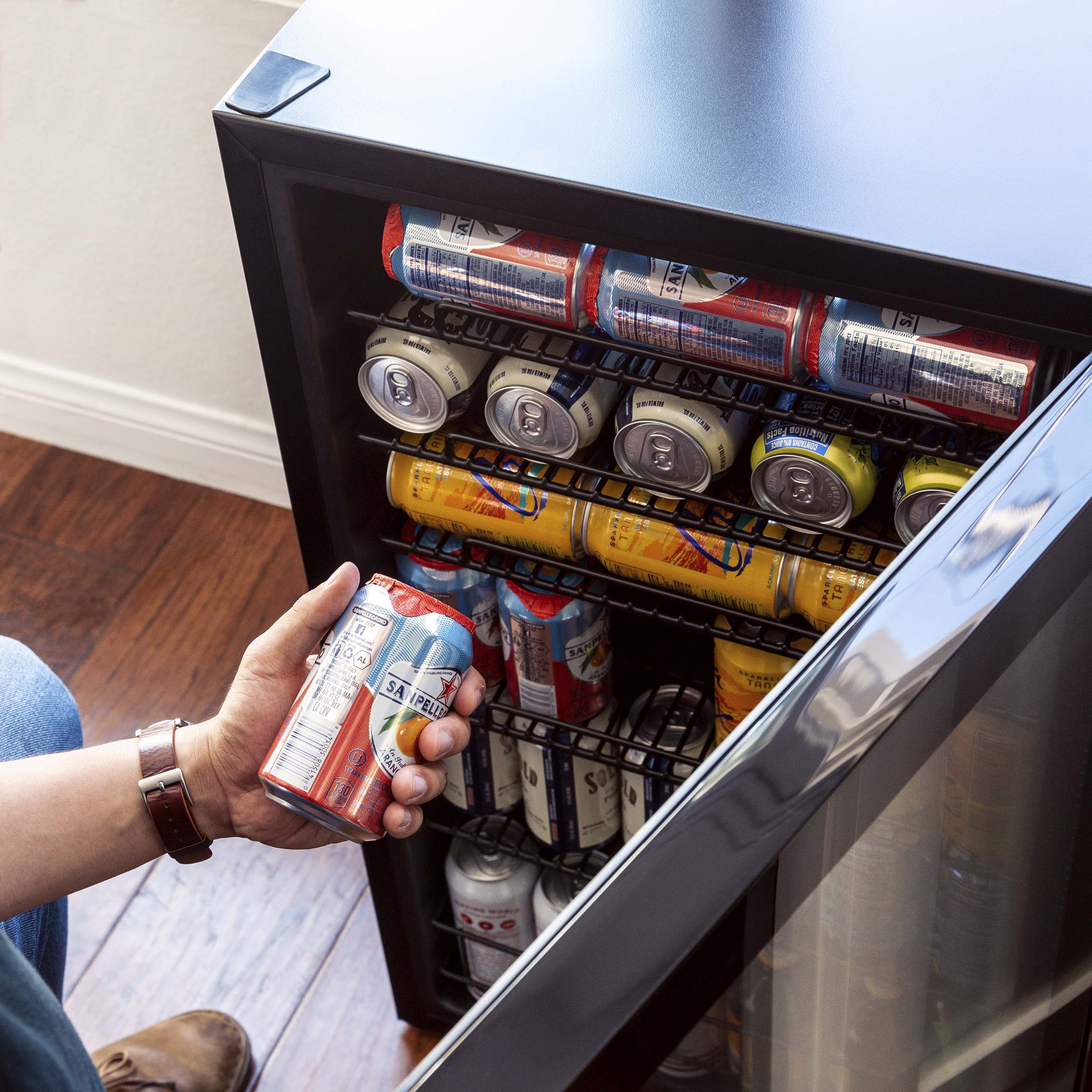 The image shows a person holding a can of beer from a refrigerator. The refrigerator is filled with various canned beverages, including a total of 13 cans. The person is likely in the process of selecting a drink from the refrigerator.