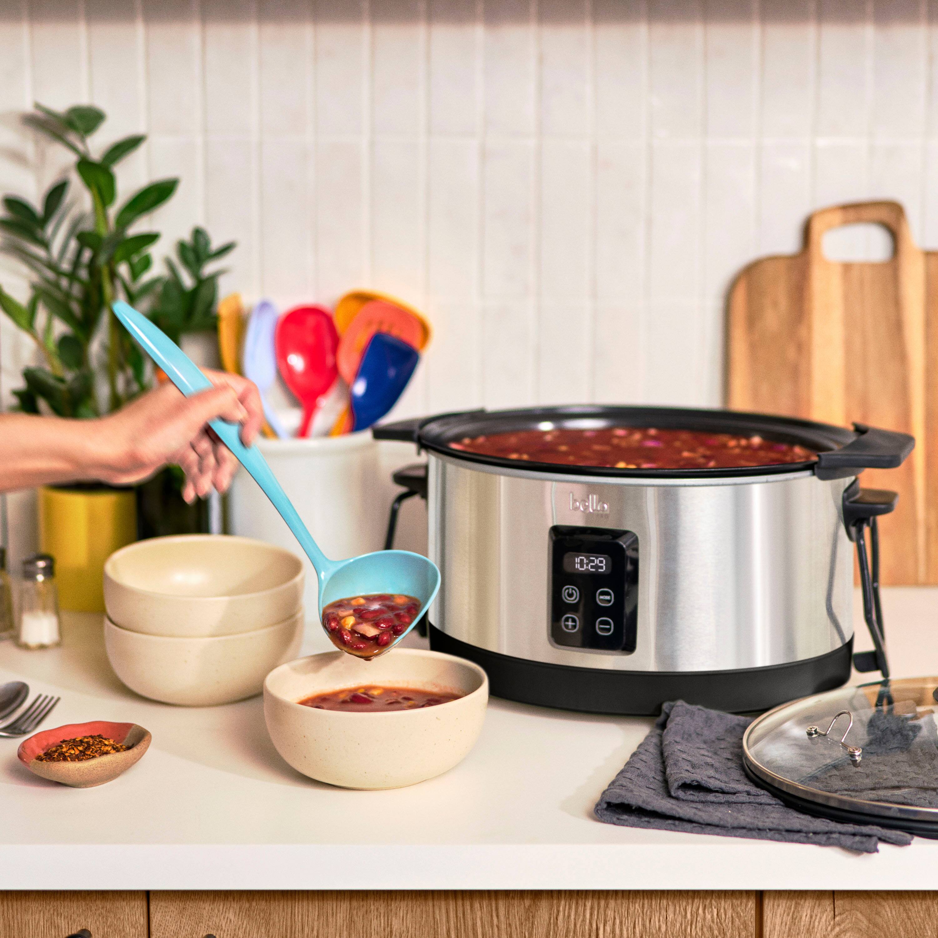 The image shows a person using a blue ladle to scoop food from a large pot on a stove. The pot is filled with red liquid, and the person is carefully serving the food into a bowl. The scene takes place in a kitchen, where various utensils and items are present.

There are multiple spoons in the image, with one near the person and others placed around the kitchen. A fork can also be seen in the scene. Additionally, there are two bowls, one near the person and another further away. A potted plant is visible in the background, adding a touch of greenery to the kitchen.