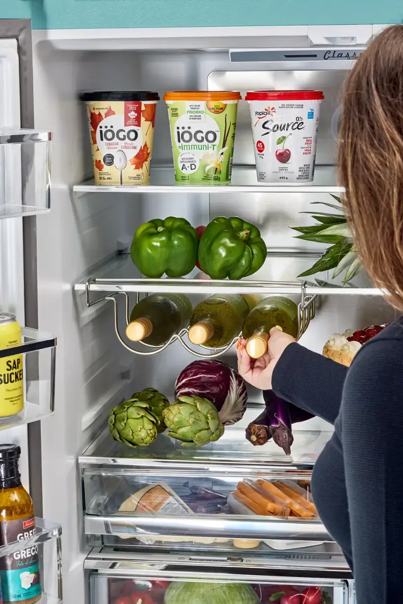 The text on the image is in French, so I cannot provide a corrected version in English. However, I can provide a general description of the contents of the refrigerator.

The refrigerator is filled with various food items, including a few bottles of drinks, a bowl, and a bunch of broccoli. There are also some fruits and vegetables, such as apples and carrots, stored in the refrigerator. The person in the image is looking at the food items, possibly deciding what to eat or preparing a meal.