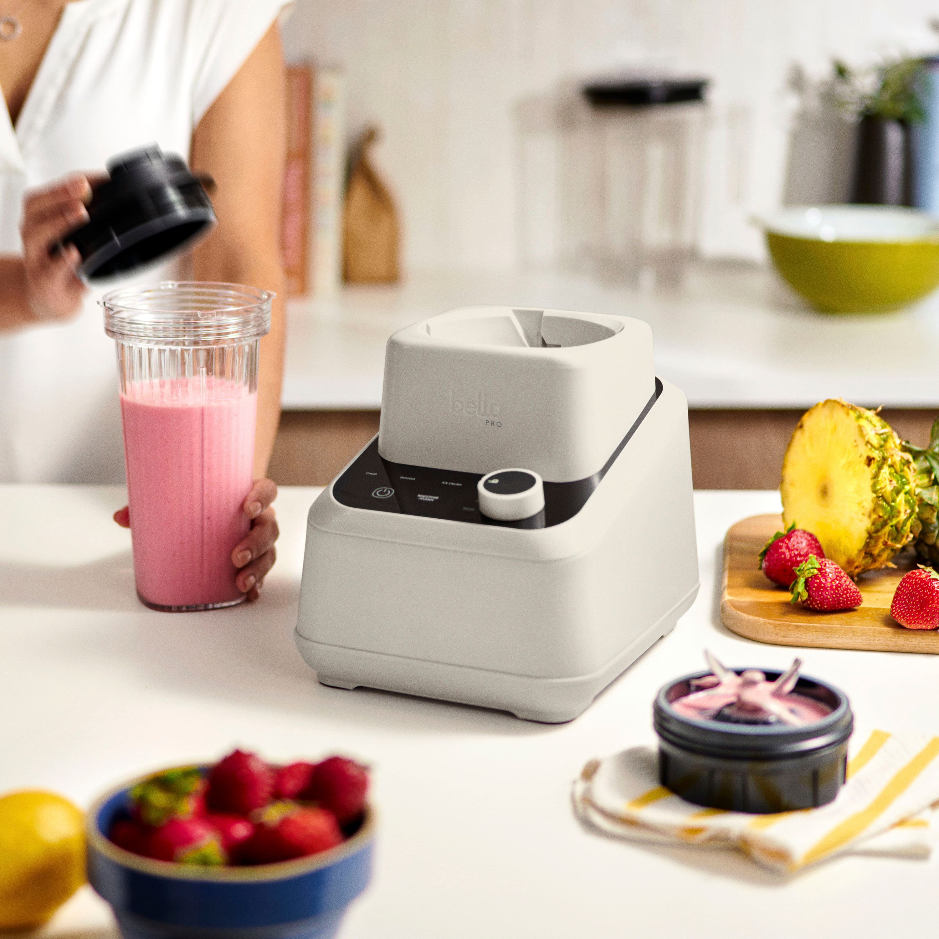 The image shows a woman standing in a kitchen, preparing a meal. She is holding a blender and a bowl of fruit, which includes strawberries and bananas. There are also other bowls and a cup on the countertop. The woman is using a blender to make a smoothie, and the scene suggests that she is in the process of preparing a healthy meal.