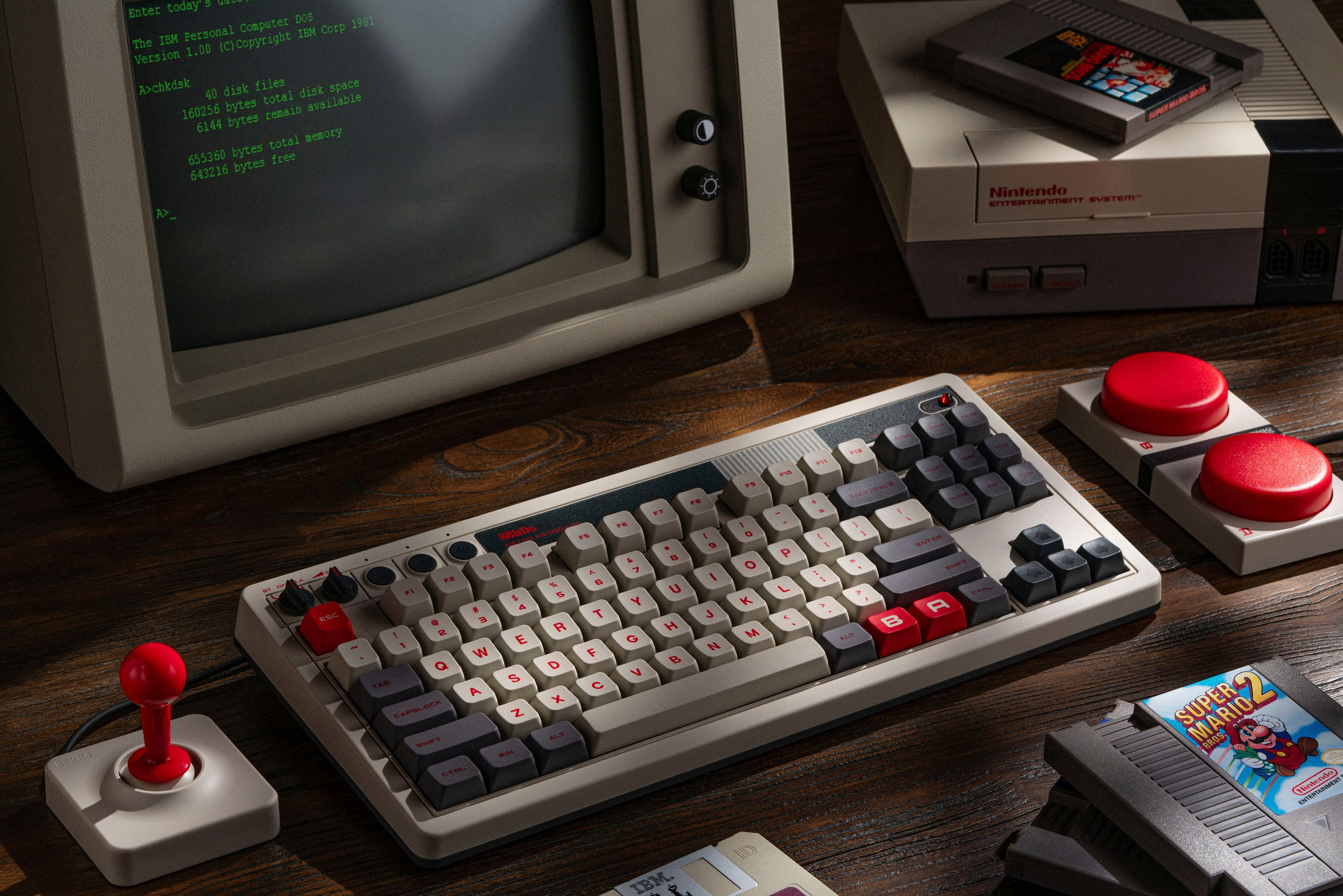 A computer keyboard from 1991 is sitting on a table next to a Nintendo Entertainment System (NES) console. The keyboard is an IBM PC keyboard, and it is accompanied by a Super Mario Bros. game cartridge for the NES. The scene also includes a computer monitor and a TV, both displaying information. The image is a black and white photo, giving it a vintage feel.