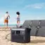 The image features a beach scene with a man and a woman walking along the shore. There is a solar panel or solar panel charger on the sand, and a portable power station or generator nearby. The man and woman are walking past the solar panel charger, which is placed on the beach.
