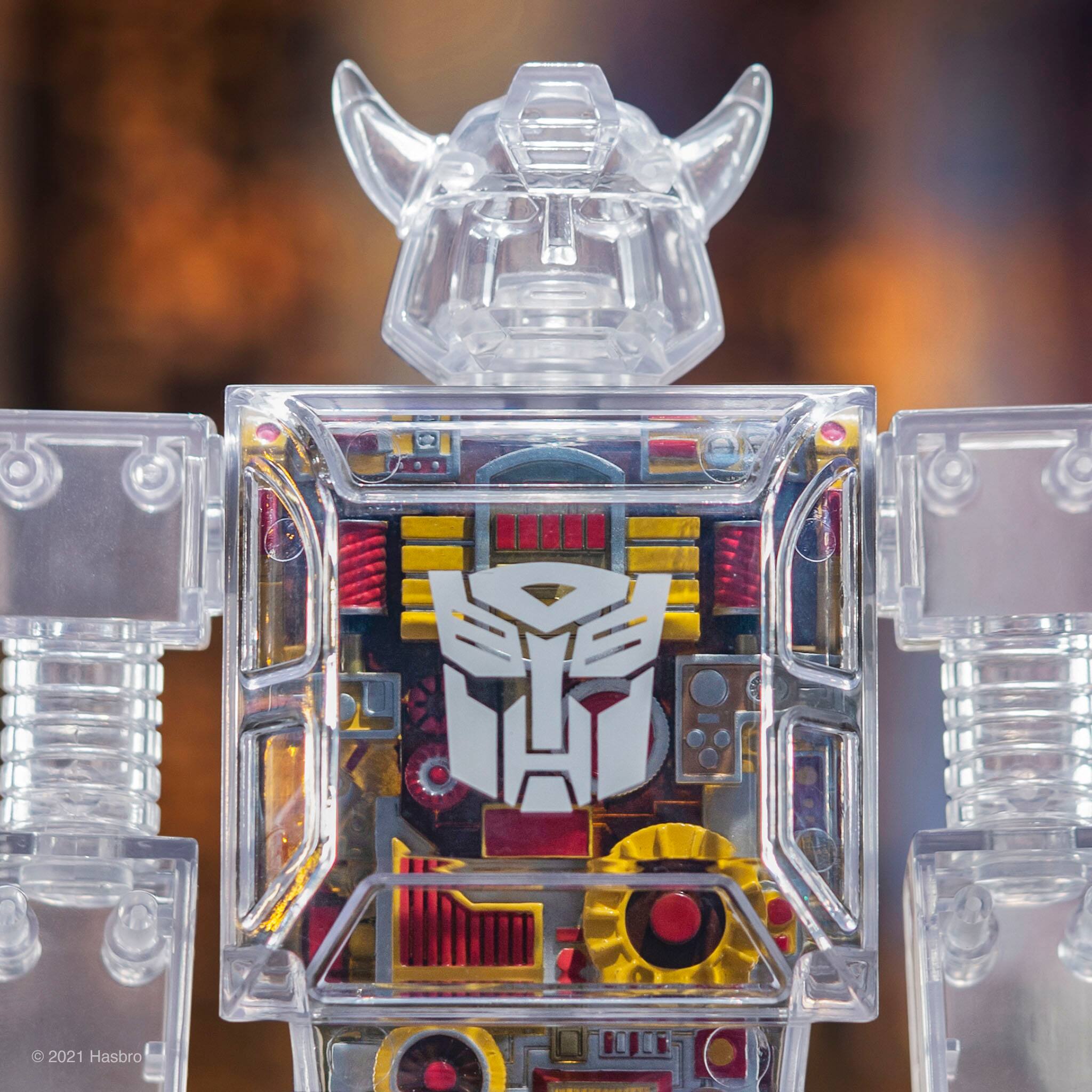 The image features a robot figurine with a face on it. The robot is made of plastic and has a clear body, allowing the internal components to be visible. The robot's face is decorated with a red and white design, and it appears to be a Transformer. The robot is standing on a surface, possibly a table or a shelf.