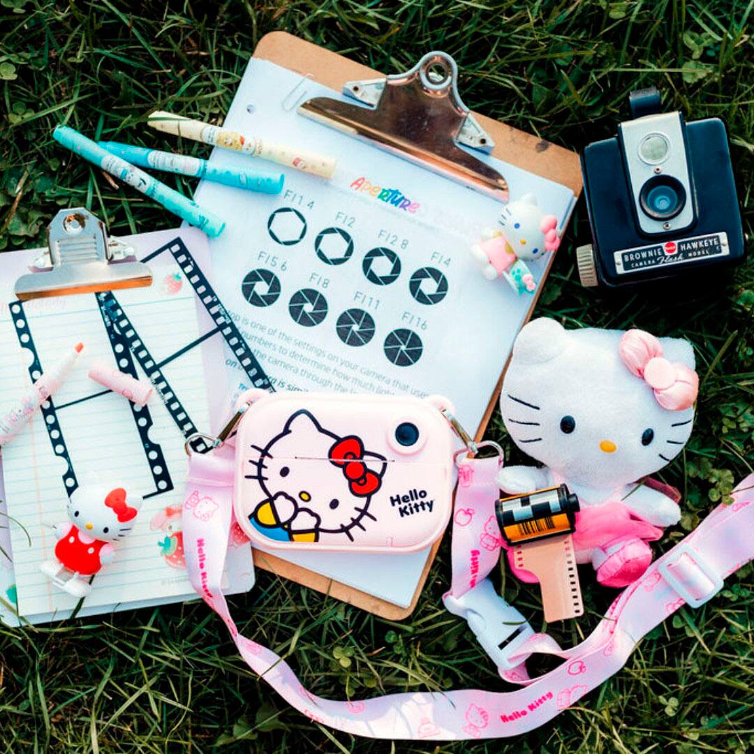 The image features a Hello Kitty lunch box, a Hello Kitty wallet, and a Hello Kitty camera. There is also a clipboard with a list of apertures, a Hello Kitty keychain, and a Hello Kitty watch. The items are arranged on a grassy field, creating a fun and playful scene.