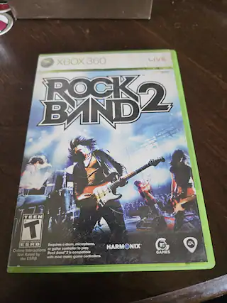 The text on the image is:
"XBOX 360 LIVE
ROCK BAND 2
TEEN
T
CONTENT RATED BY ESRB
Online Interactions Not Rated by the ESRB
Requires a drum, microphone, or guitar controller to play. Rock Band 2 is compatible with most music game controllers.
HARMONIX GAMES
EA"
Corrected and grouped text:
"XBOX 360 LIVE
ROCK BAND 2
TEEN
T
CONTENT RATED BY ESRB
Online Interactions Not Rated by the ESRB
Requires a drum, microphone, or guitar controller to play. Rock Band 2 is compatible with most music game controllers.
HARMONIX GAMES
EA" - T (Teen 13+)