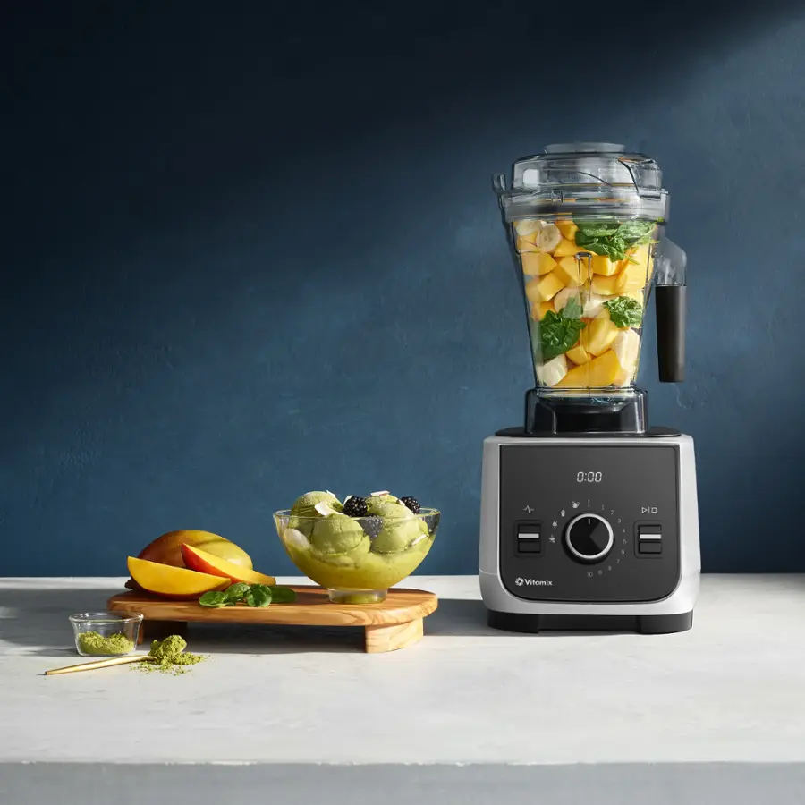 The image shows a table with a blender and a bowl of fruit. The blender is filled with fruit, and there are various fruits, including apples and oranges, placed around the table. The blender is set to zero, indicating that it is ready to blend the fruits. The table is white, and the overall scene suggests a healthy and refreshing atmosphere.
