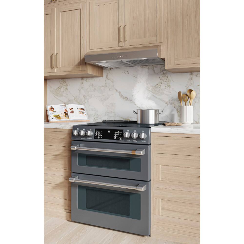 The image shows a kitchen with a stove top oven. The oven has a stainless steel finish and is located under a ventilation hood. The kitchen also features a marble backsplash and wooden cabinets. There are several spoons placed on the countertop, and a book is visible on the counter as well. The overall scene is well-lit, creating a warm and inviting atmosphere.