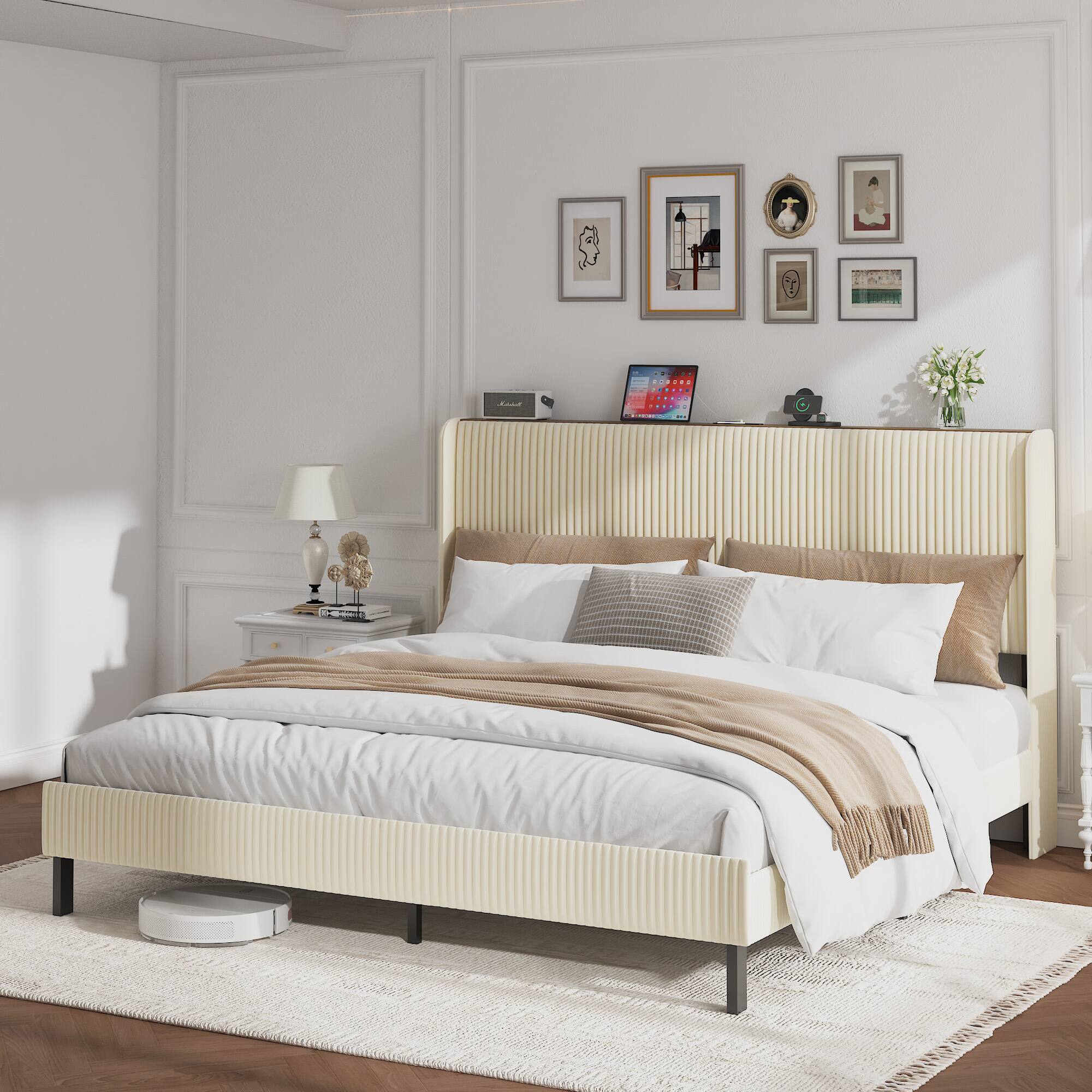 The image shows a neatly arranged bedroom with a modern and minimalist design. The room features a large bed with a beige upholstered headboard and white bedding, complemented by beige and brown throw blankets. On the wall above the bed, there are several framed artworks and photographs. To the left of the bed, there is a bedside table with a lamp, a small clock, and a few books. On the right side, there is a small shelf with a laptop, a digital clock, and a vase with white flowers. The room has white walls with decorative molding, and the floor is covered with a light-colored rug. The overall aesthetic is clean, bright, and inviting.