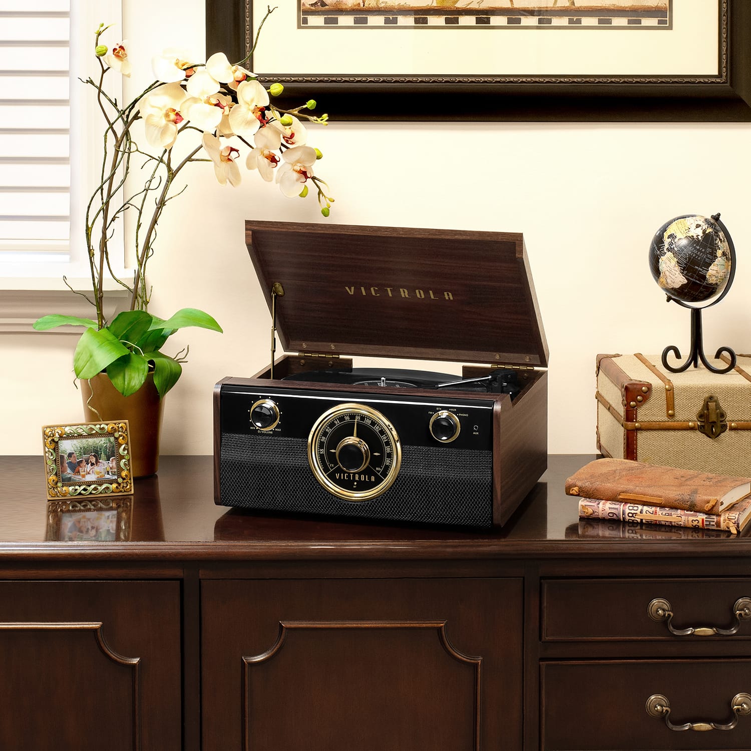 The image features a vintage Victrola record player sitting on a wooden desk. The record player is open, revealing its intricate design and the turntable inside. The desk also has a few other items on it, including a potted plant, a vase, and a book. The overall scene gives a nostalgic and cozy atmosphere, reminiscent of a bygone era.