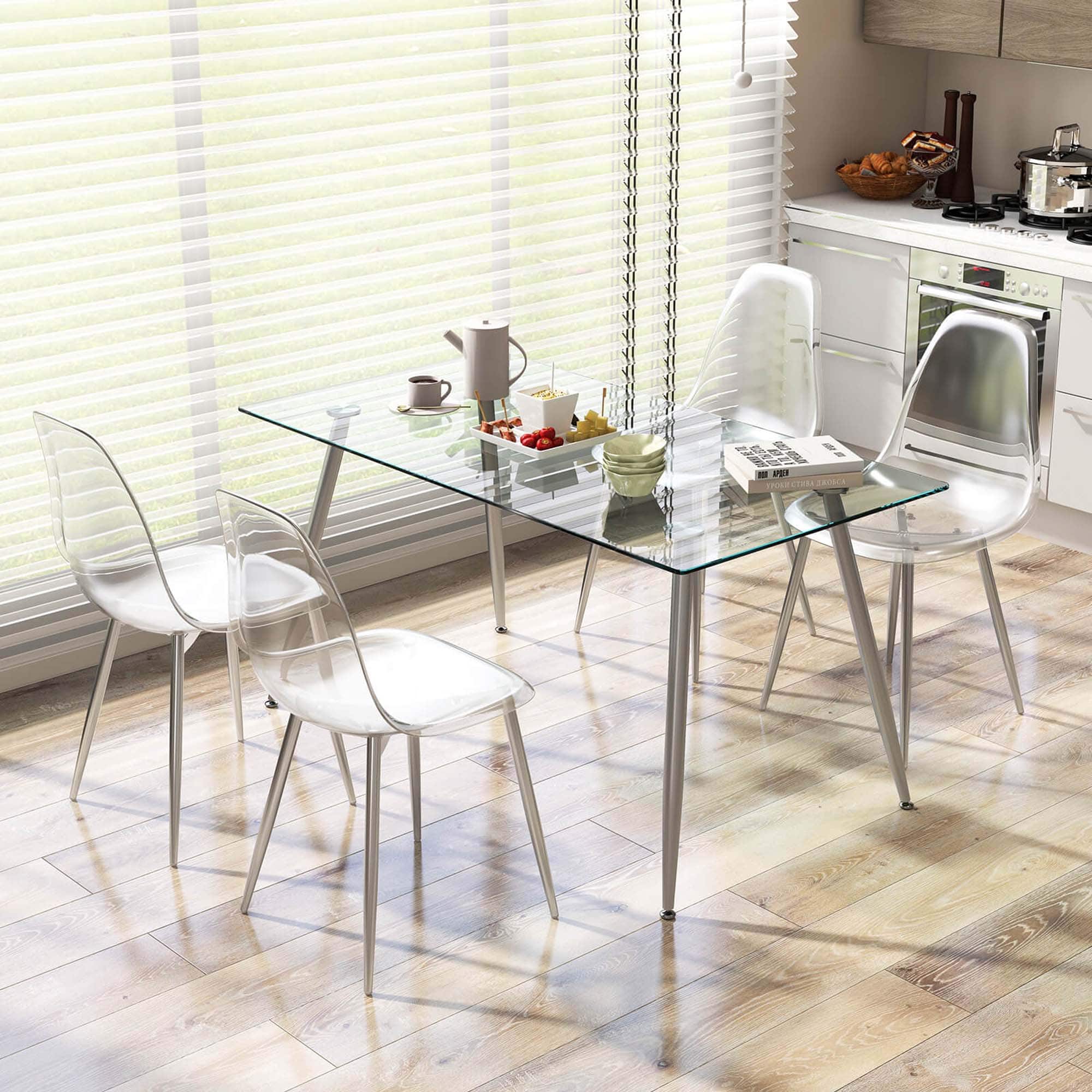 The image features a glass dining table with four chairs surrounding it. The table is set with various items, including cups, bowls, and a book. There is also a sink and an oven in the background, indicating that the scene takes place in a kitchen. The table is positioned in front of a window, allowing natural light to illuminate the space.