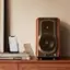 The image features a wooden table with a speaker and a cell phone on it. There is also a book and a remote control nearby. The scene suggests a comfortable living space where someone can enjoy music, read a book, or watch TV. The presence of the cell phone and remote control indicates that the person might be using these devices for entertainment or communication purposes. The book on the table adds a touch of leisure and relaxation to the scene, while the speaker and cell phone represent modern technology and convenience. Overall, the image portrays a cozy and well-equipped living space.
