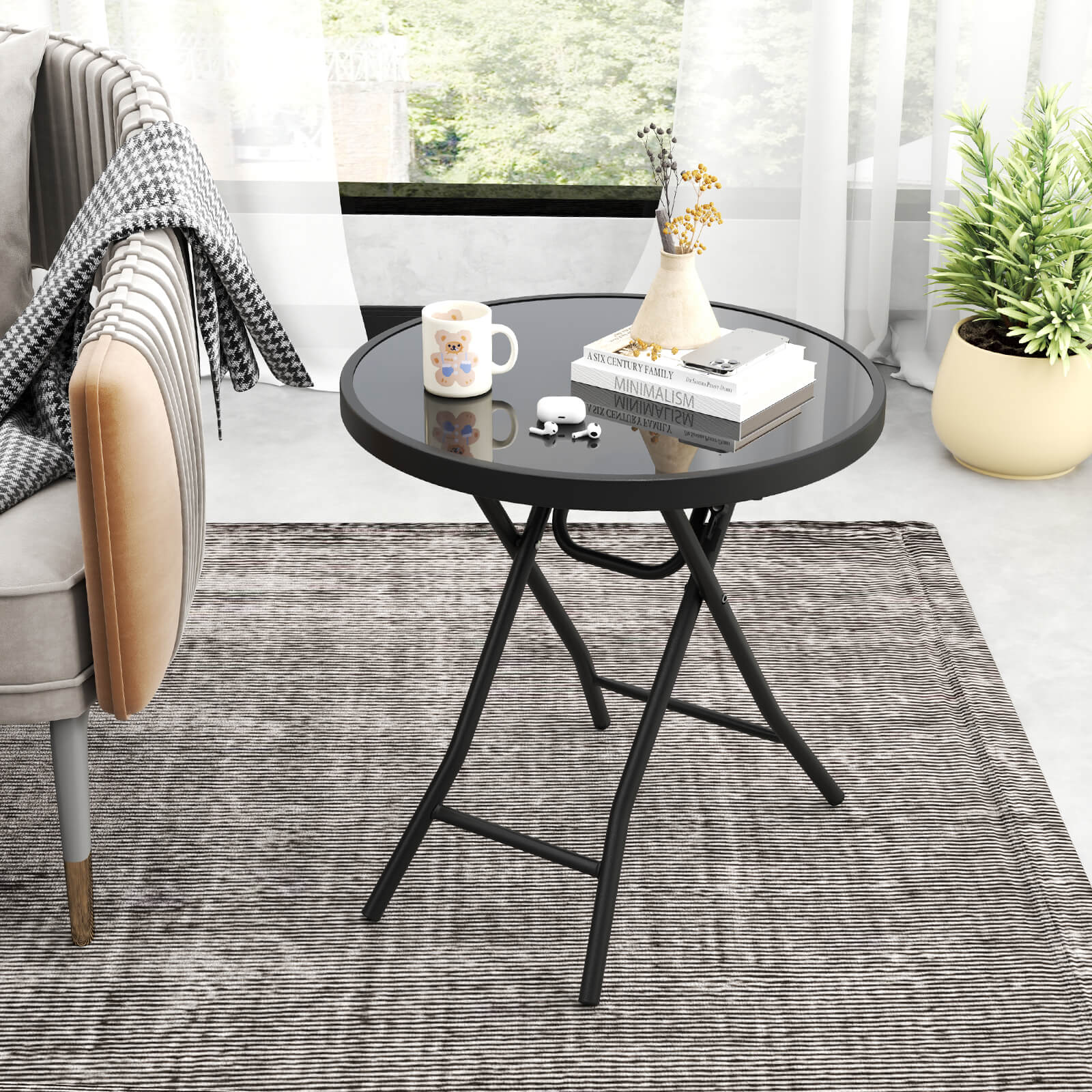The image features a black coffee table with a white vase on top. The table is placed on a rug in front of a window. There is a chair nearby, and a potted plant can be seen in the background. The table also has a book and a cup on it. The scene appears to be a cozy living space with a focus on minimalism.