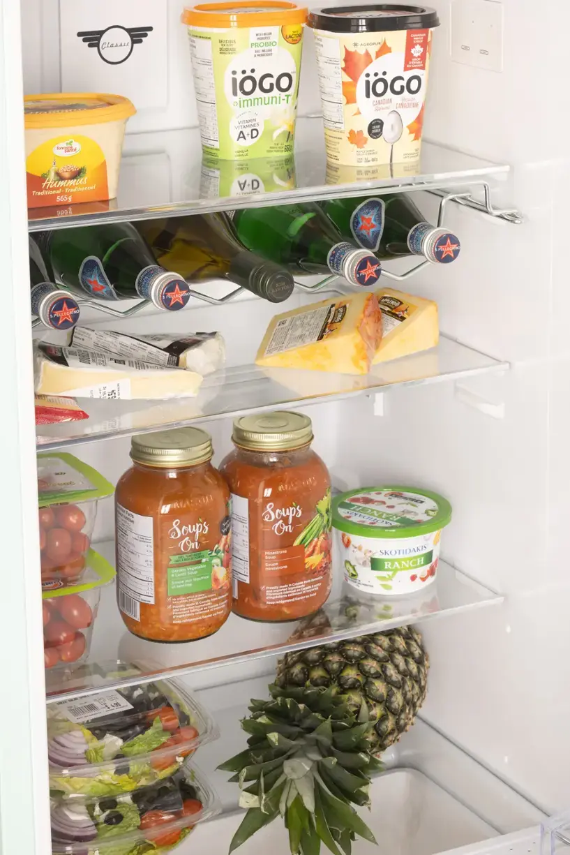 The image shows a refrigerator filled with various food items, including a pineapple, soup, and other items. The text on the image reads:

"Classic n Hummus Traditionnel Traditional 565g 009 & - . -- - - LACTS 1 BLE - hes . - PROBIO as -U   |  .. .. -. 1 1N iGO - T% eimmuni-T - 2 .. . JOUTI . .. VITAMIN VITAMINES A-D - - I - . - Sa  - -  CTS - - - A-D .  -   SOPUR 10 -  - - iGO CANADIAN ScAir Statens CANADIEME -  -  - .   - C aT - - - - T al -   PALACAIO PELLEGI AIM CRR 1 LLICT - Tum - Soups On   . - - - Ganson Vegetabre & LASEO Su - Mgumes SouDe . an ANS -  - - SA - - a S - I - -  rdr - PAlACie  1 A Fretil saer - - Soup's On Minestrone Smar SOuET - - - marT - Pa  A ncu   mie - - da - PRn  -   Ntr - - HONYY SKOTIDAKIS RANCH I - 99"