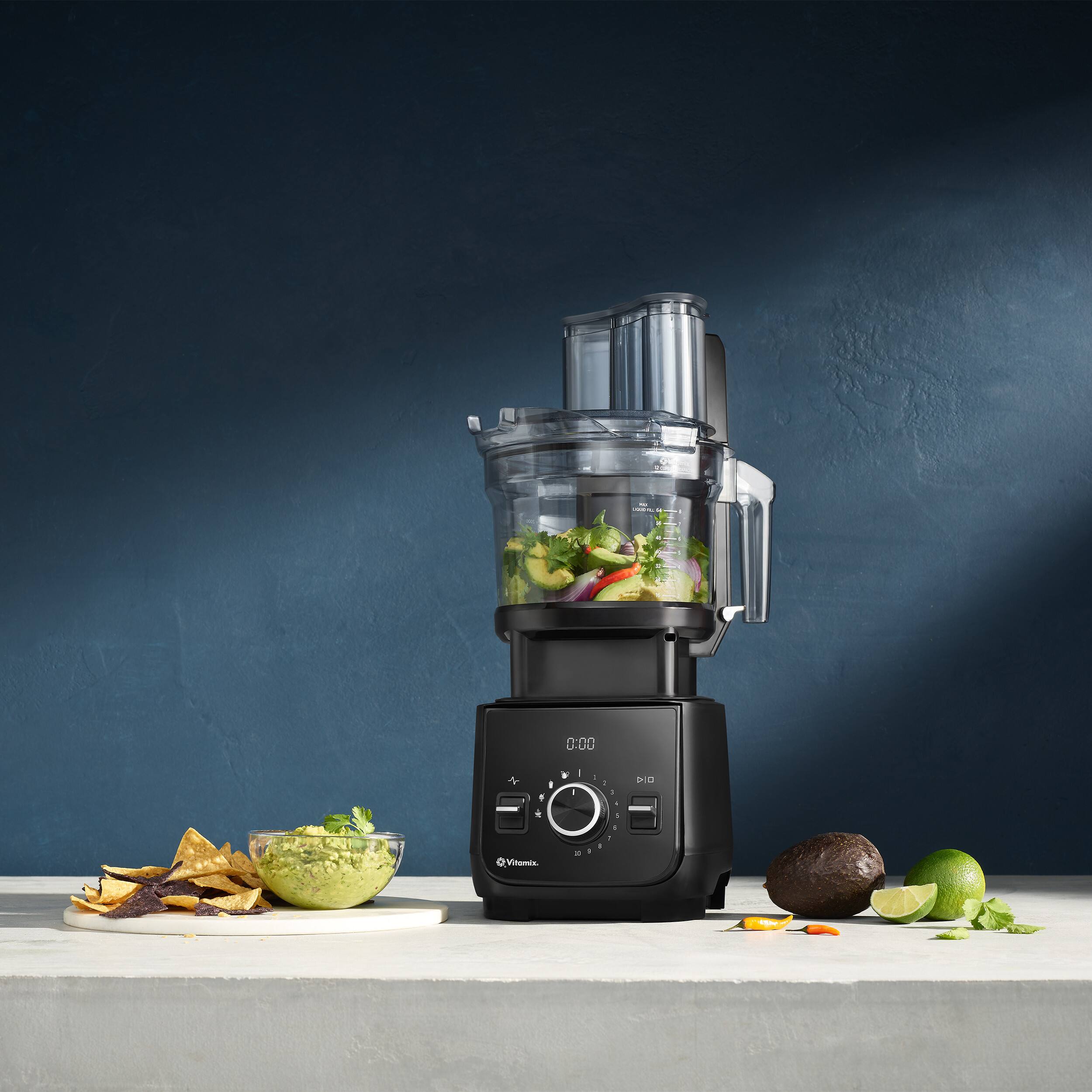 The image shows a blender on a counter with a bowl of guacamole and a plate of chips. The blender is filled with a green mixture, possibly guacamole or a smoothie. There are also a few carrots and a lime placed nearby. The scene suggests a healthy snack or meal preparation.