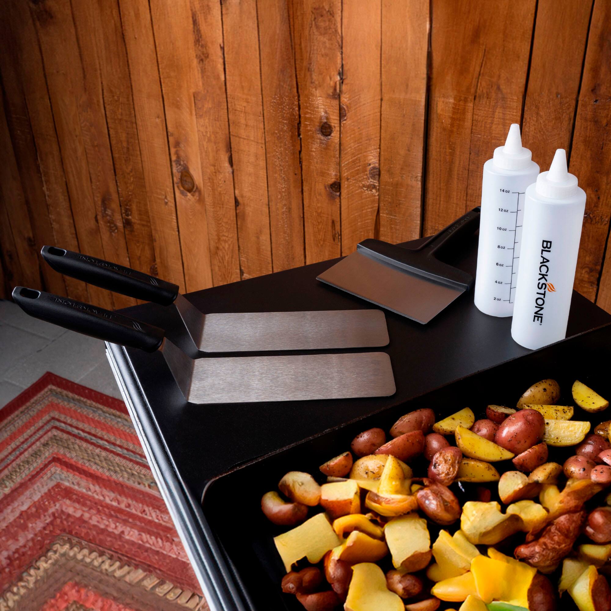 The image features a wooden table with a cutting board and various food items, including potatoes, apples, and bananas. There are also two bottles of Blackstone seasoning on the table. The table is set up for food preparation, with the cutting board and food items ready for use.