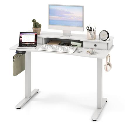 The image shows a modern, white standing desk with a minimalist design. On the desk, there is a computer monitor displaying a serene beach scene, a laptop with a calendar open, a keyboard, a mouse, a small potted cactus, a cup of coffee with a spoon, a pen holder with pens, and a round wall clock. There is also a hanging bag on the side of the desk.