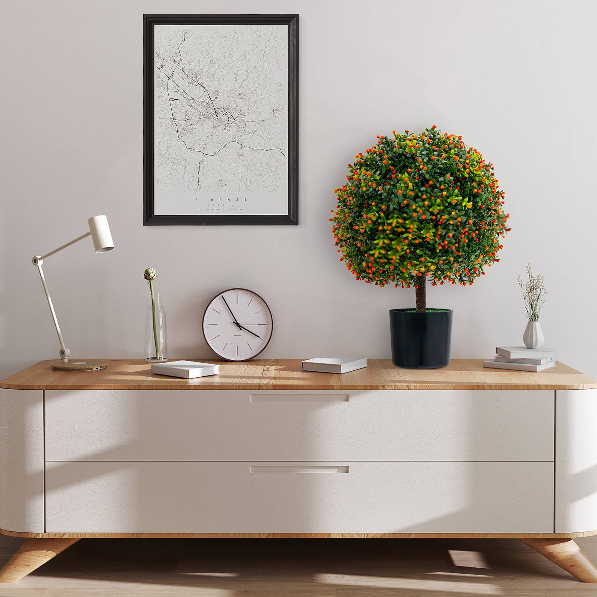The image shows a neatly arranged wooden console table against a light-colored wall. On the table, there is a modern white desk lamp on the left, a round wall clock with a white face and black hands, and a small stack of books. In the center, there is a potted plant with lush green leaves and orange berries. To the right of the plant, there is a small white vase with dried flowers. Above the table, a framed map of Florence is hung on the wall. The table has a light-colored drawer front with wooden handles, and the floor is made of light wood.