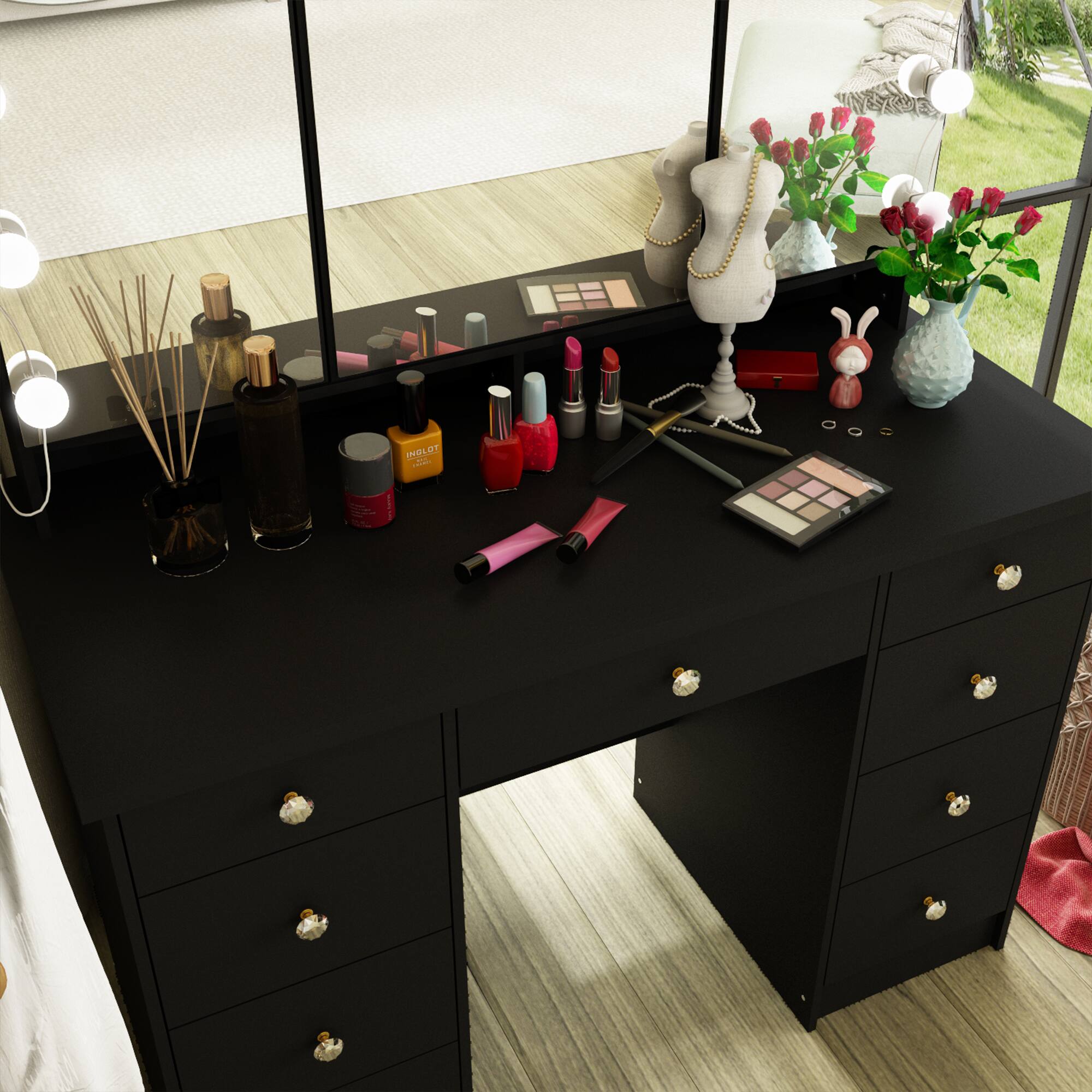 The image shows a black vanity table with a mirror, featuring various beauty products and accessories. On the table, there are:

- Several bottles of nail polish in different colors.
- A makeup palette.
- A lipstick.
- A small vase with flowers.
- A diffuser with reed sticks.
- A small decorative bunny.
- A jewelry stand with a necklace.
- A few other small containers and makeup items.

The vanity has drawers with decorative handles.